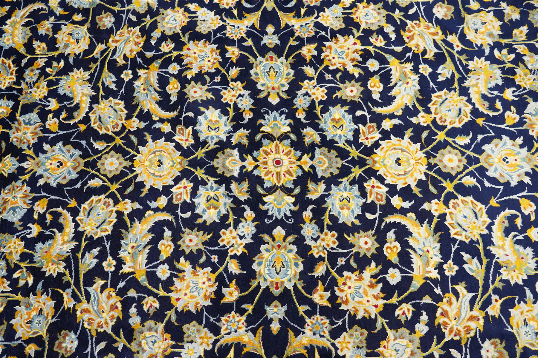 Central Persian Kashan hand-knotted carpet, deep indigo field with a densely all-over arrangement of scrolling floral vines, palmettes, and flowering sprays, in soft ivory, pale blue, and warm gold tones, principal ivory border with a continuous meandering vine of stylized palmettes, and floral motifs, flanked by multiple narrow guard borders, with cotton fringe ends 