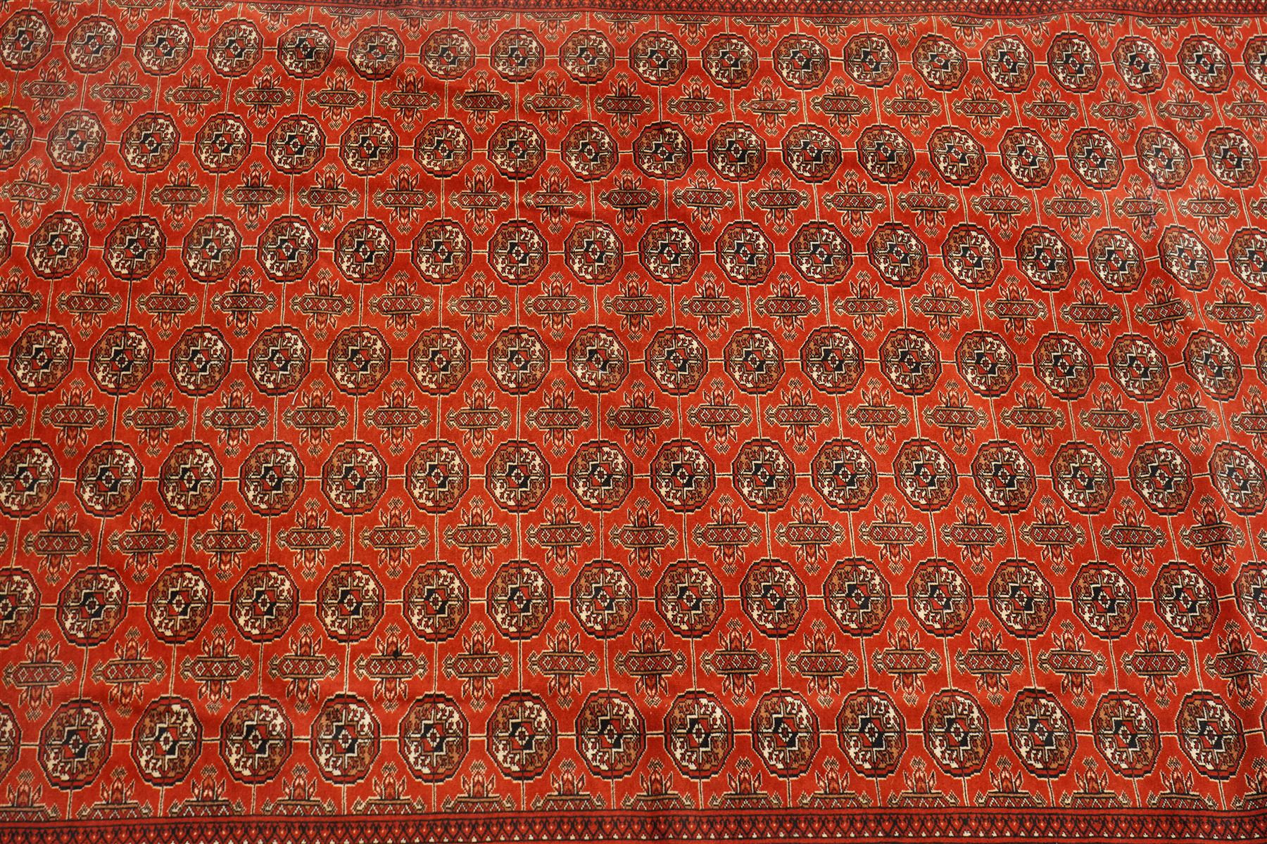 Turkmen hand-knotted carpet, madder red field, with an all-over design of row of Tekke guls, each enclosing stylized cruciform and geometric devices in tones of ivory, dark brown, and indigo, principal border of stylized geometric motifs, flanked by guard border with angular latch hook geometric designs, with cotton fringe ends 