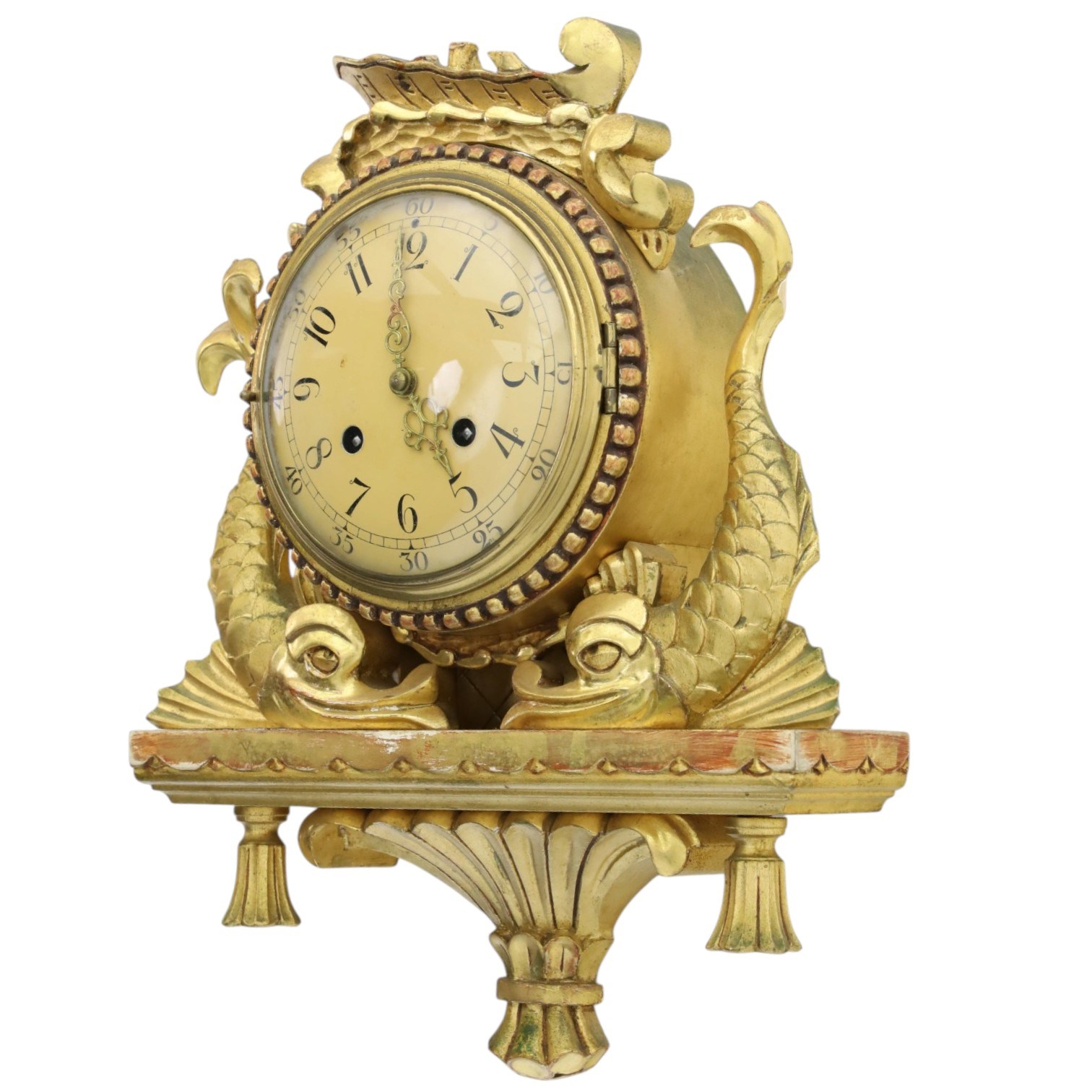 20th century Continental giltwood wall clock, circular dial with Arabic numerals and pierced gilt hands, within a carved case surmounted by a scrolling crest and flanked by stylised dolphins, above a shaped shelf with tassel pendants and a foliate terminal, pendulum missing
