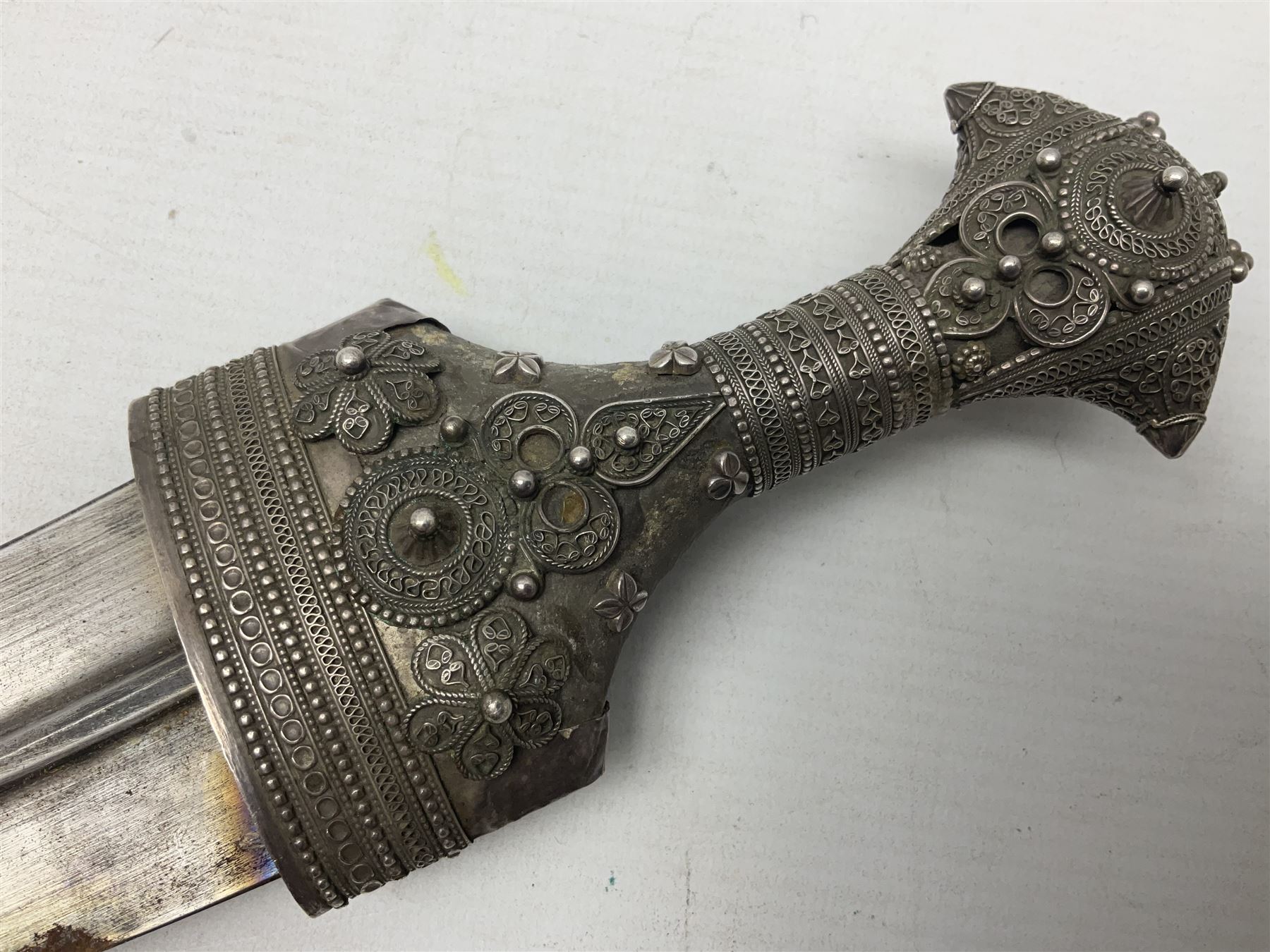 Omani Khanjar dagger, the 19cm curved blade with pronounced medial rib to either side, waisted hilt with dome shaped pommel and silver covered horn grip decorated with filigree bands, floral motifs and beadwork; in an intricately filigree decorated silver and leather scabbard with silver rings woven into the belt mount and fitted with original silver thread-work belt L34cm overall belt length 94cm