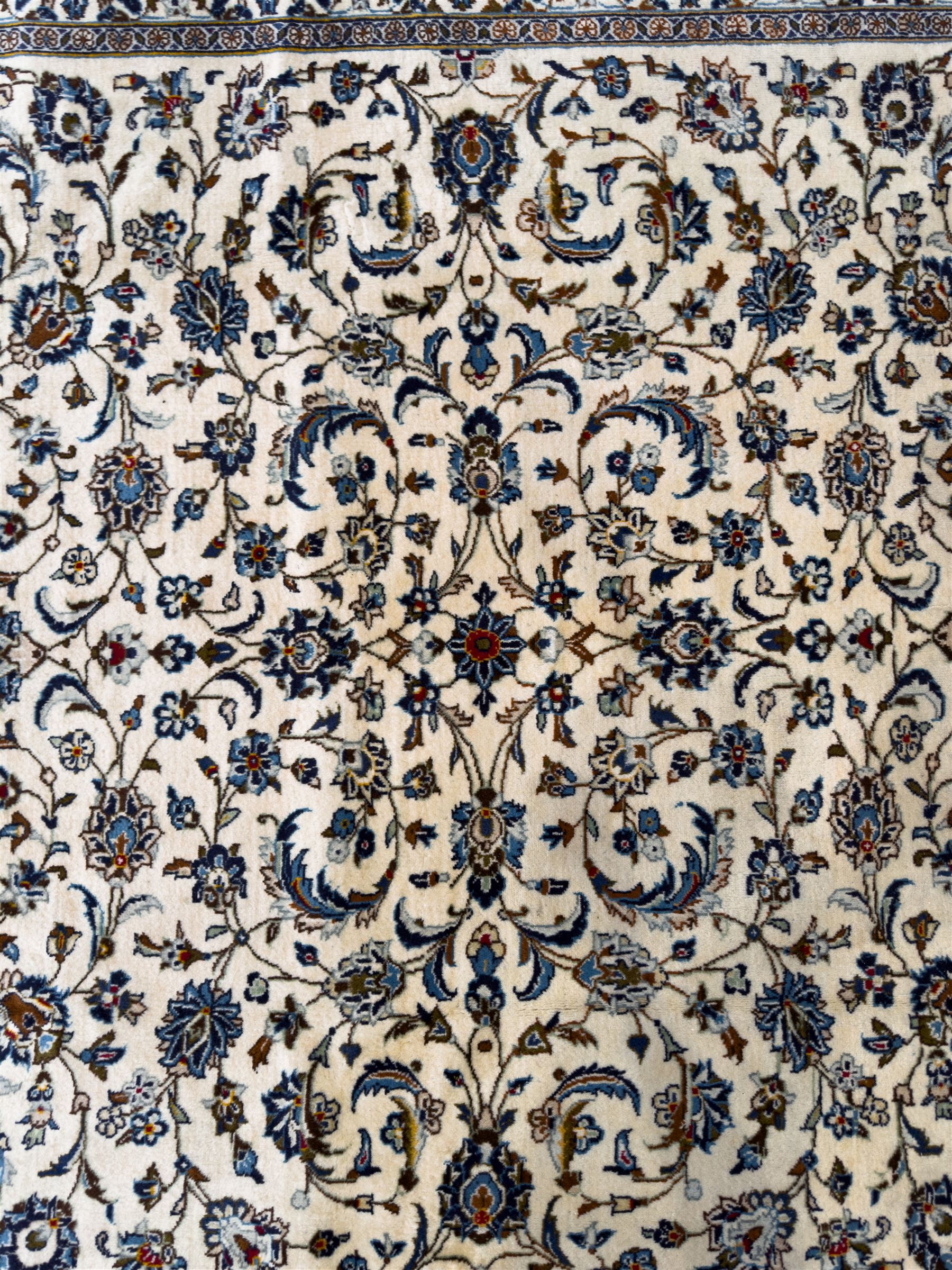 Central Persian Kashan hand-knotted carpet, ivory field with an all-over design of scrolling vines, lotus palmettes, and rosettes in shades of blue and brown within a blue ground border of palmettes and arabesques between multiple guard stripes, 
348 cm x 246cm