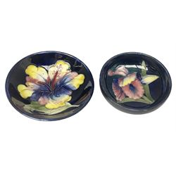 Moocroft small footed circular dish decorated in the Hibiscus pattern upon cobalt blue ground, with Queen Mary paper label beneath, together with a smaller example decorated in the Orchid pattern with impressed and printed W.Moorcroft marks beneath, largest, D9cm, (2)