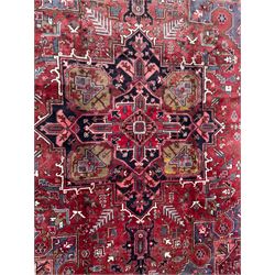 Northwestern Persian Heriz hand-knotted carpet, madder red field with a bold stepped medallion in indigo, ivory, and ivory, surrounded by angular floral motifs and stylized leaves, and the spandrels set within a dark border of rosettes and angular vine motifs, 335cmx252cm