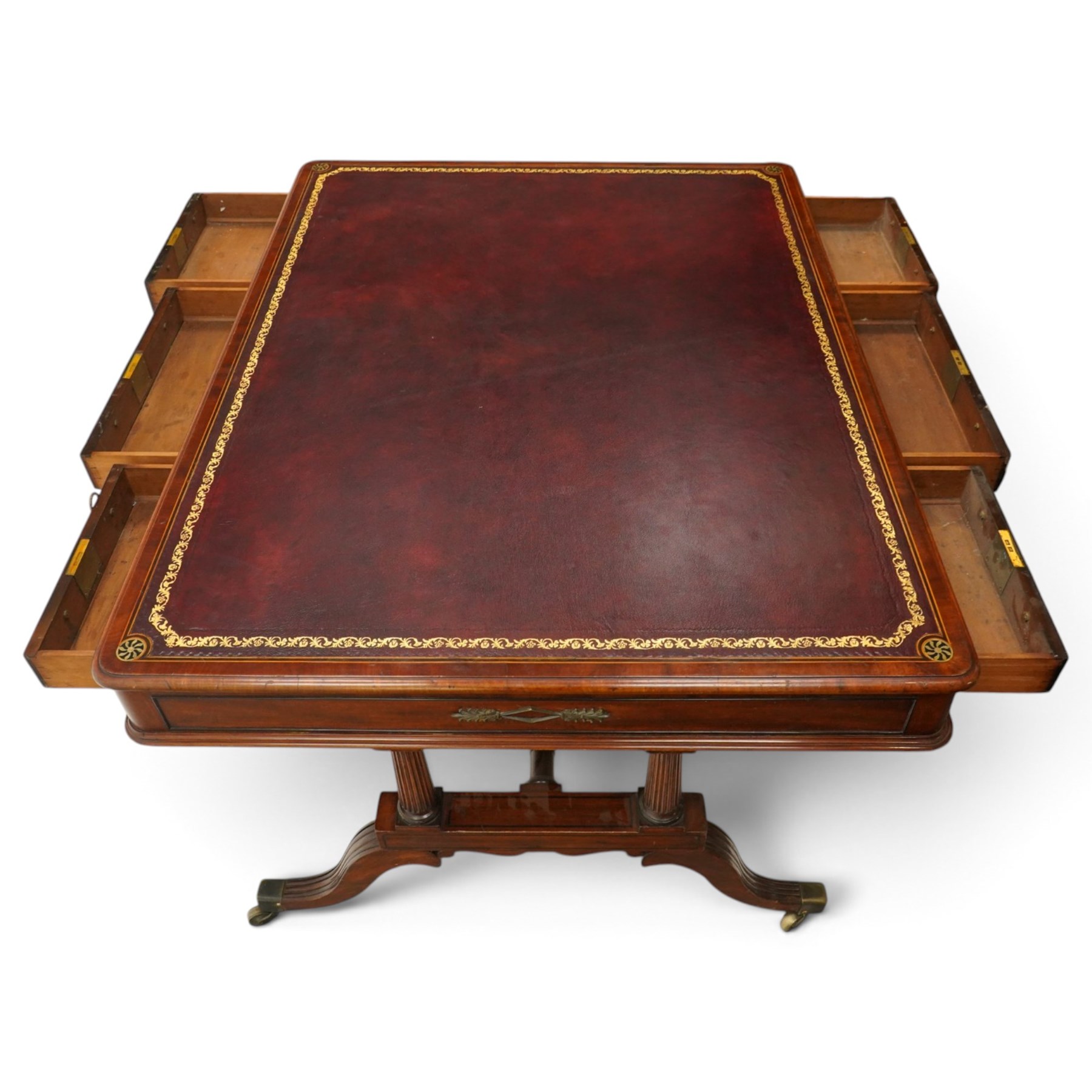 Regency inlaid mahogany and brass mounted library table, the rectangular top inset with gilt tooled burgundy leather writing surface, enclosed by  crossbanding and boxwood stringing with roundel corner motifs, above three cock-beaded frieze drawers to either side fitted with brass ring handles and brass escutcheons, the end friezes centred by applied brass foliate mounts, raised on twin reeded column supports over rectangular platform ends, united by a ring-turned stretcher, the reeded sabre legs terminating in brass caps and castors, one drawer bearing ivorine label 'Spillman & Co., St Martins Lane, London'