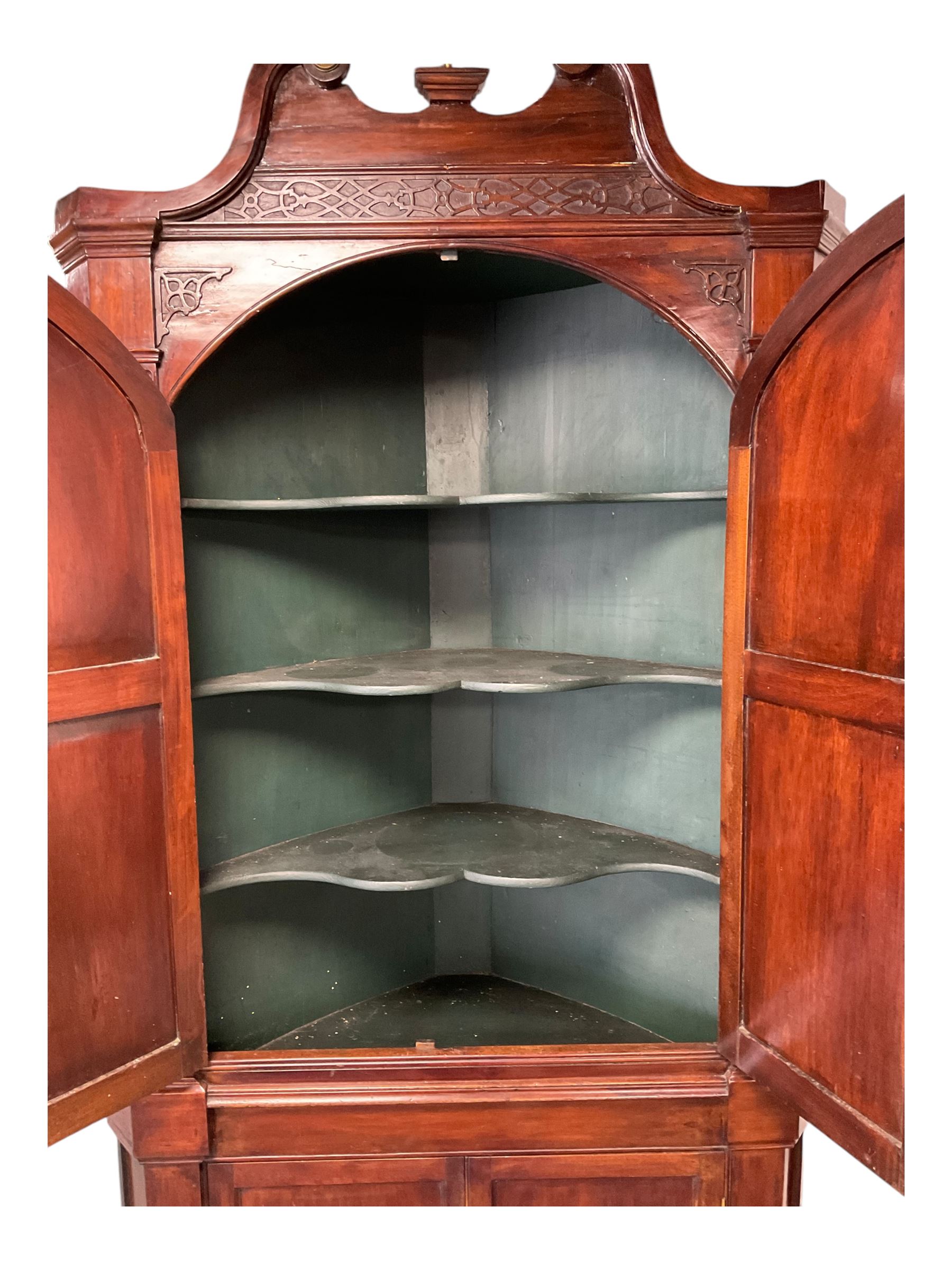 George III mahogany corner cabinet, the scrolled pediment surmounted by a brass finial and carved fretwork frieze, with fluted uprights flanking arched double doors over a painted interior fitted with shaped shelves, above a lower cupboard with twin panelled doors, on plinth base