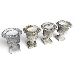 Set of four weathered composite stone urns of campana form, the everted leaf-moulded rolled rim over a body decorated with scrolling fruiting grapevines in relief, over a gadrooned underbelly and waisted socle, lobed collar on square base