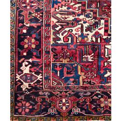Northwestern Persian Heriz hand-knotted carpet, red madder field with a bold stepped medallion in indigo, ivory, and terracotta, flanked by angular floral motifs and stylized leaves, the spandrels set within a dark indigo border of rosettes and angular vine motifs between narrow guard stripes, 340cm x 245cm 