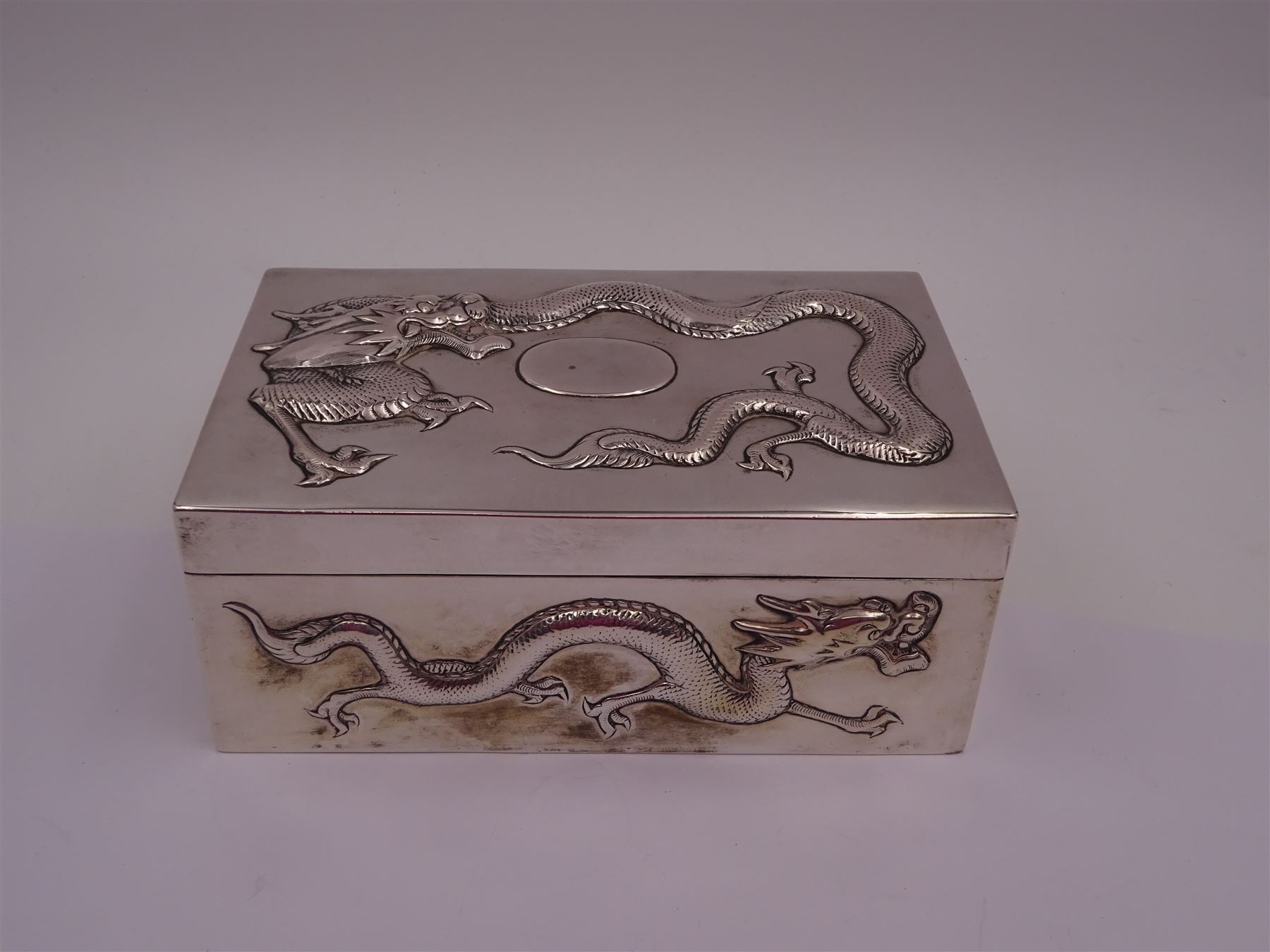 Late 19th century/early 20th century Chinese export silver mounted cigarette box, of rectangular form, decorated in relief with dragons, the hinged cover with similar dragon decoration and central blank oval cartouche, opening to reveal a softwood lined compartmentalised interior, stamped beneath with character mark and KMS for maker Kwong Man Shing, H6.3cm, W15.5cm, D9.5cm