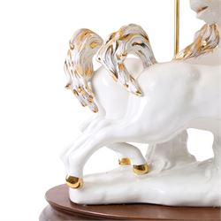 Italian-style ceramic table-lamp featuring two white running horses with gold accents, with cream tasselled lampshade, upon a wooden base, approx. H60cm W40cm