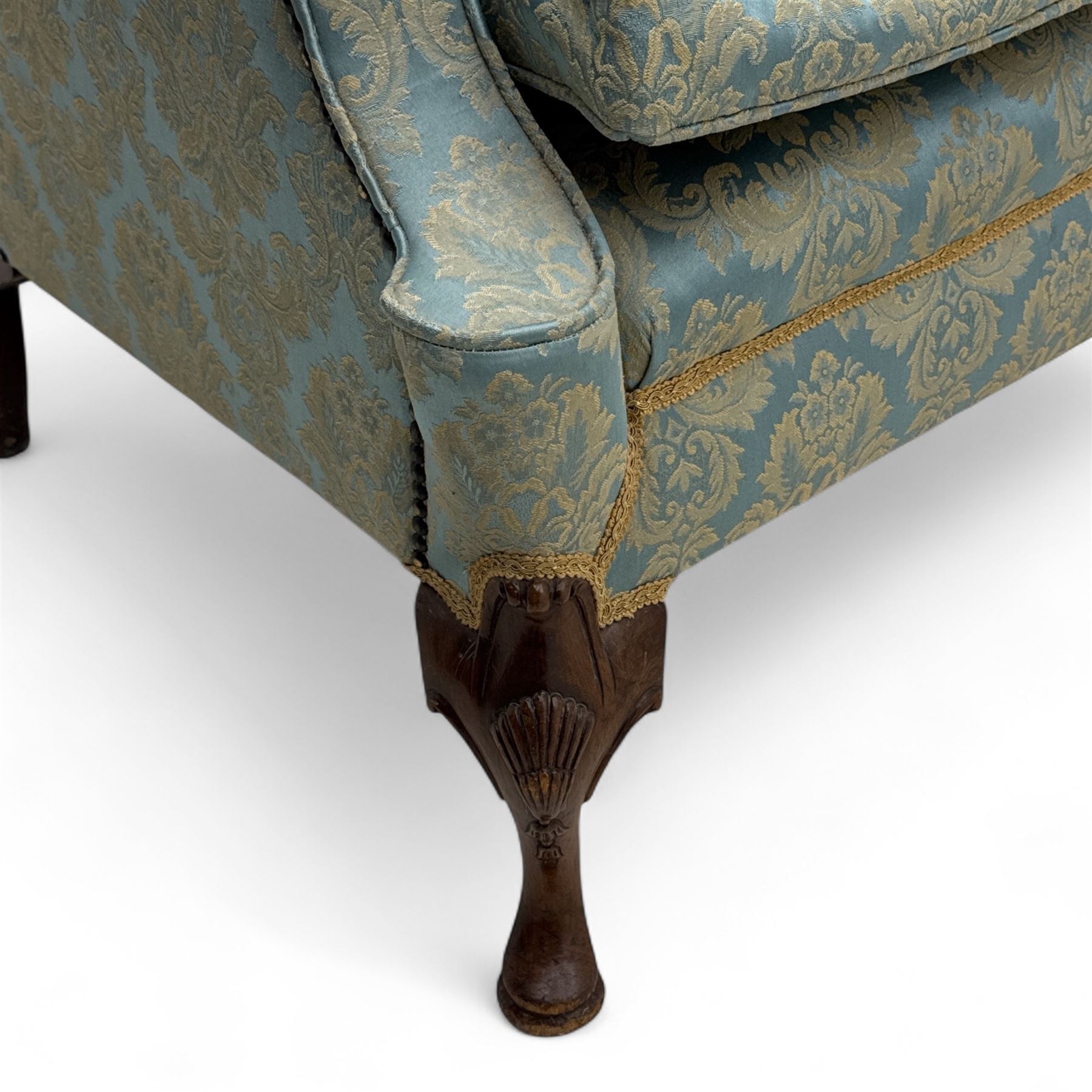 Georgian design carved walnut framed wingback armchair, upholstered in pale blue and gilt floral damask, with shaped loose seat cushion and rolled arms, raised on scallop and cartouche carved front cabriole supports terminating to pad feet
