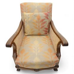 Pair of mid-20th century mahogany framed bergère armchairs, each with carved scroll arms and caned side panels, floral upholstered loose cushions, raised on cabriole front supports