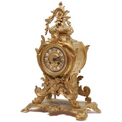 French-Late 19th century 8-day gilt brass rococo style mantel clock, c1890, waisted case  decorated with scroll work, leaves and musical instruments, resting on an attached plinth with splayed feet, dial within a glazed brass bezel with a gilt repoussé centre, porcelain cartouche Roman numerals and steel fleur-di-Lis hands, with a twin train movement, recoil anchor escapement and countwheel strike, sounding the hours and half-hours on a bell. With a matching pair of five-light candelabra. No Pendulum or Key.