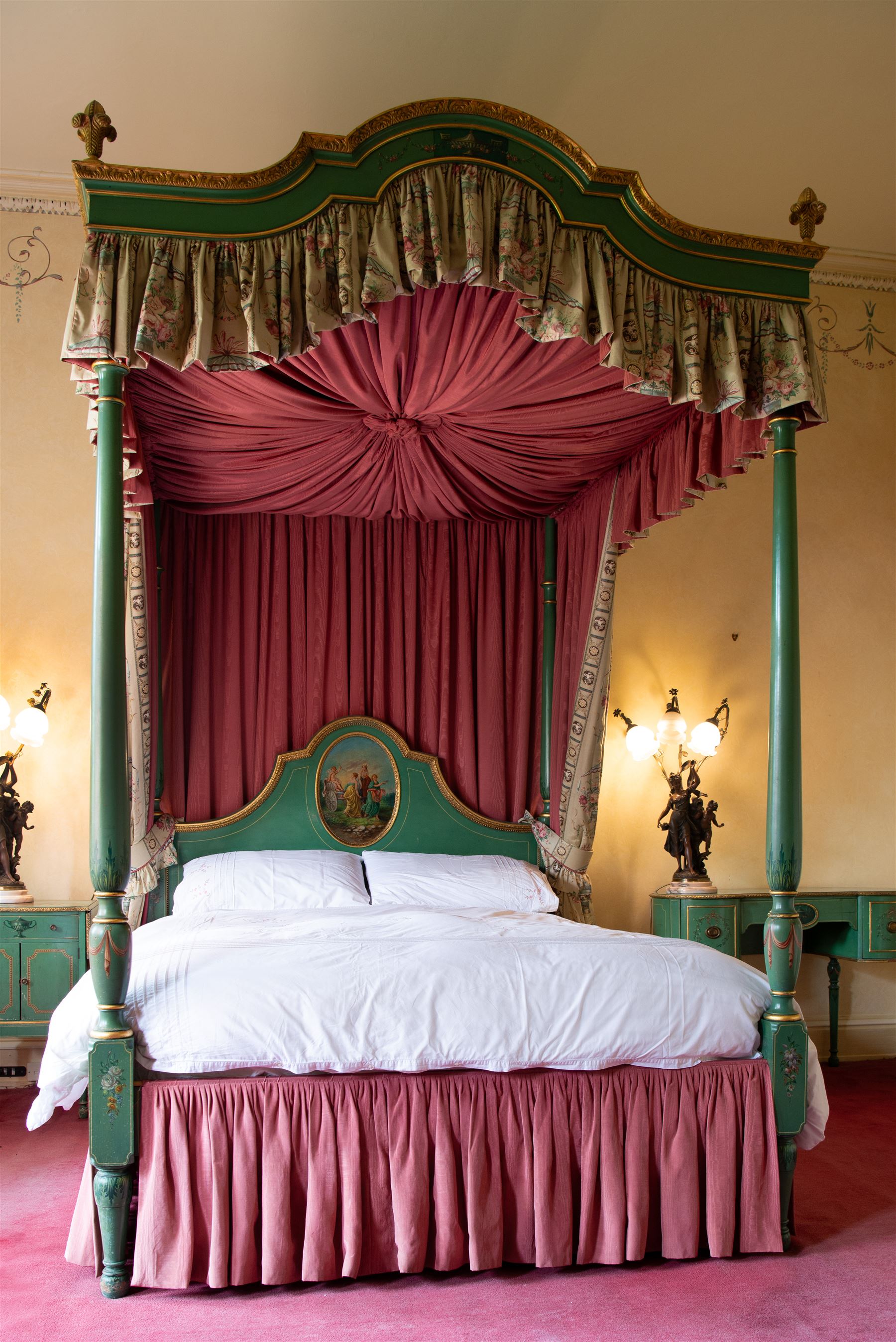 Edwardian Adam Revival green-painted and parcel gilt 5' Kingsize four poster-bed, acanthus leaf moulded canopy with curled feather finials, on four turned posts painted with acanthus leaves and draped festoons, shaped arch headboard with painted oval panel depicting maidens of love leading a chariot holding Eros, with craquelure finish, dressed in silk curtains decorated with floral bouquets 