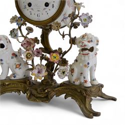 French- early 20th century 8-day Chinoiserie brass and porcelain mantel clock in the 18th century style, brass base with splayed feet profusely decorated with clusters of painted porcelain flowers and two decoratively painted Chinese foo dogs, movement enclosed in a circular porcelain movement case illustrated with birds of paradise and oriental motifs, with a white enamel dial Arabic numerals, minute markers, pierced gilt hands and a floral swag, rack striking Parisian movement striking the hours and half-hours on a bell. With a visible sunburst pendulum and key.
