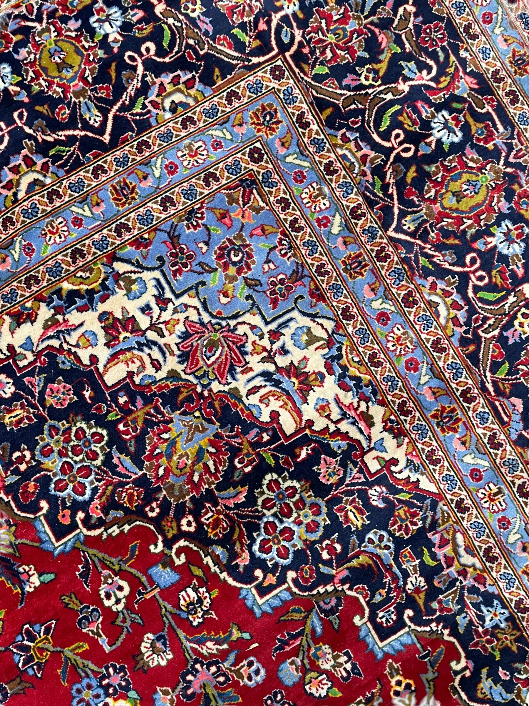 Persian Kashan crimson ground carpet, floral pattern indigo ground central medallion and matching spandrels, the field decorated densely with scrolling foliate patterns and floral motifs, the main border decorated with scrolling branches and palmettes, within floral pattern guard stripes