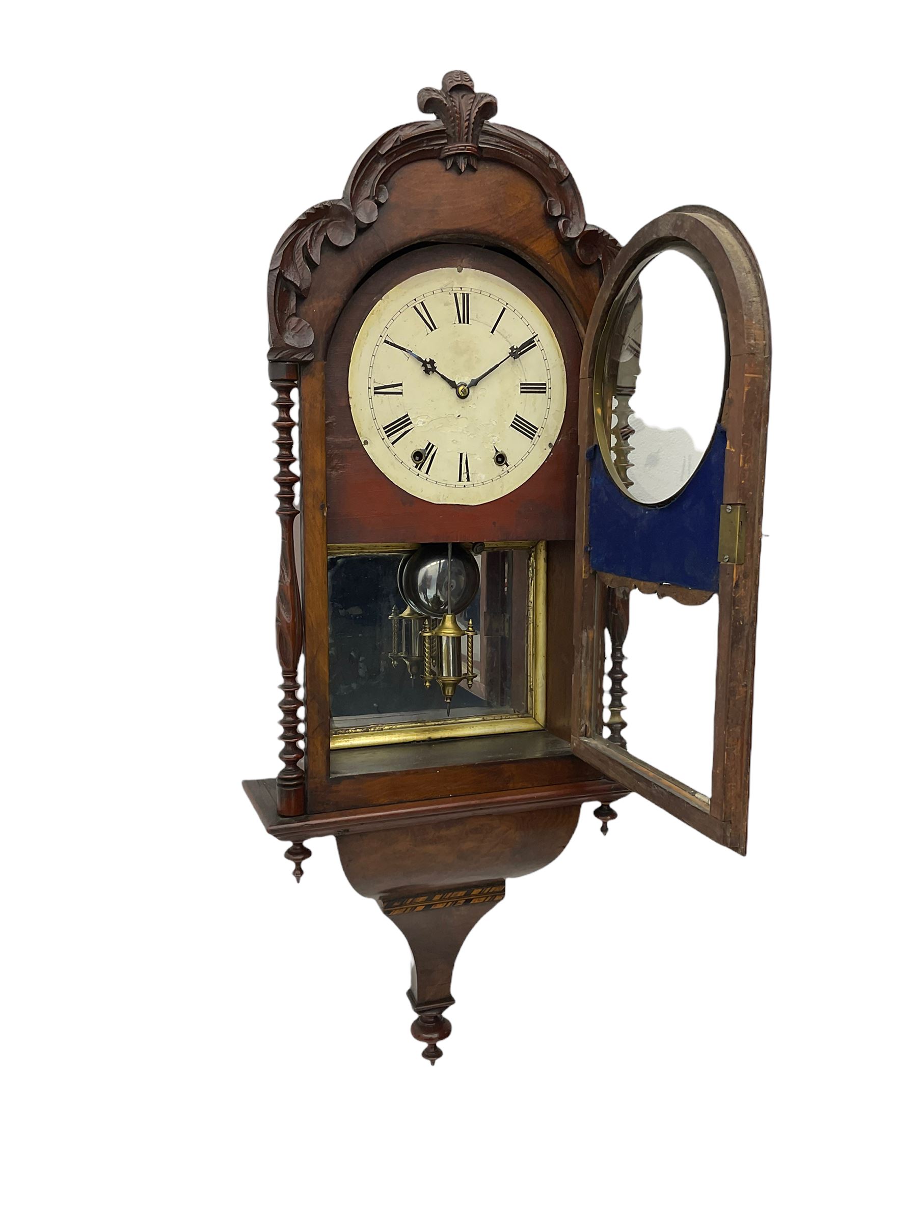 A 19thcentury American wall clock in a walnut case with crossbanding, with a carved pediment surround and arched glazed door flanked by two ring turned columns with pendants on an ogee plinth beneath, with a spring driven movement striking the hours on a bell, eight-inch painted dial with roman numerals, minute track and steel Maltese-Cross hands, with a spun brass bezel and winding collets, visible mock mercury pendulum with a reflective silvered mirror to the rear. With key and pendulum.   

