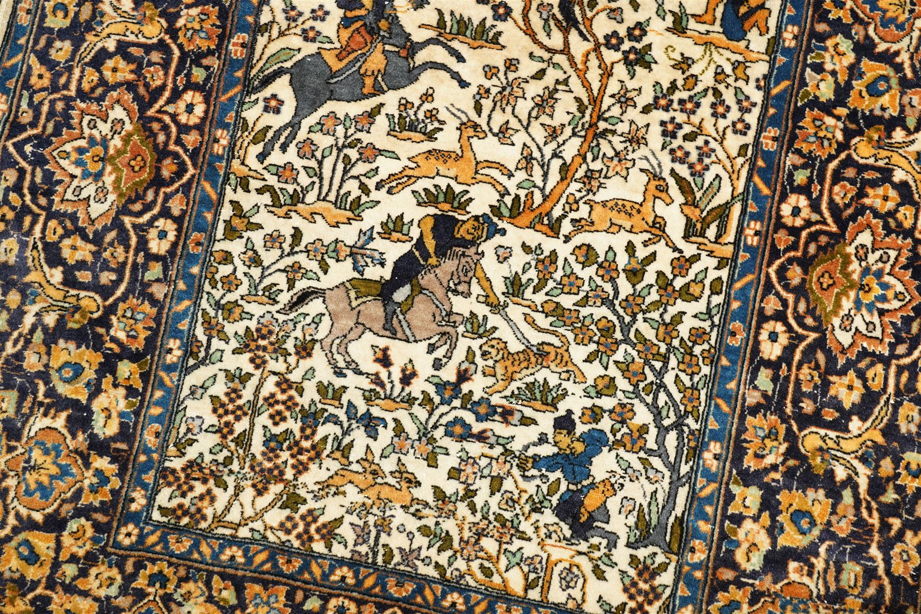 Persian pictorial rug featuring a hunting scene, ivory field with mounted hunters, animals, birds and trees, rich navy and terracotta floral border with rosettes and palmettes 