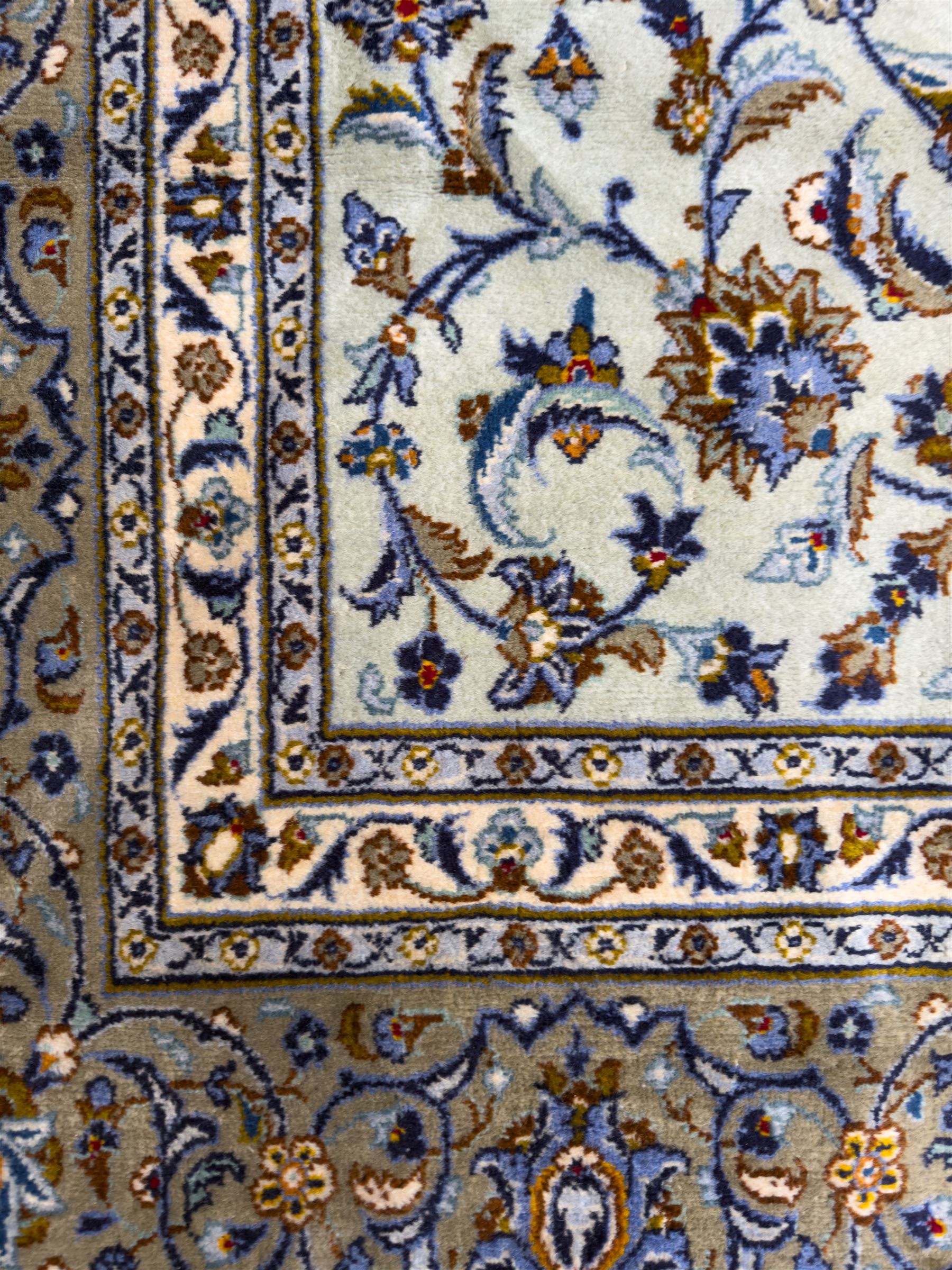 Central Persian Kashan hand-knotted carpet, the ivory ground intricately decorated with scrolling vines, palmettes, and stylised flowerheads in soft blue, gold and brown tones, enclosed by a broad indigo border with repeating floral motifs and multiple guard stripes