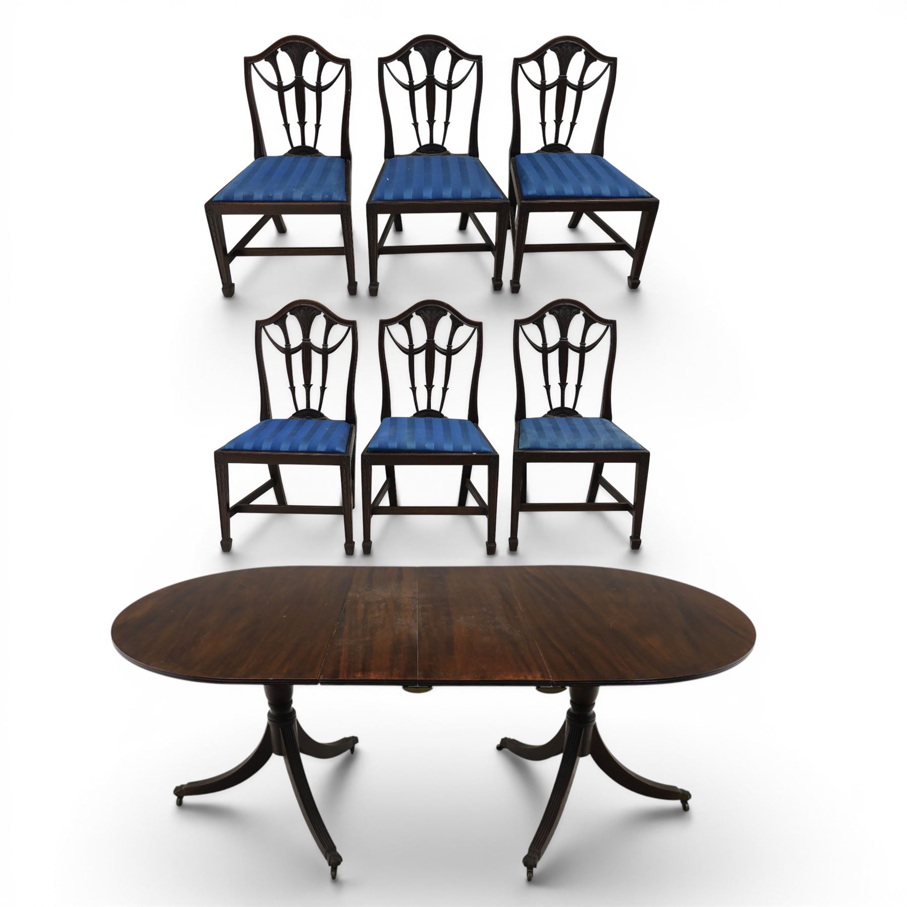 Set of six George III style mahogany dining chairs, comprising six singles with shaped top rail above pierced splay carved with feathers, with drop in stuffover seats upholstered in blue striped fabric, raised upon square tapering legs joined by stretchers, together with a mahogany twin pedestal dining table, the oval top with central leaf raised upon turned columns and splayed reeded legs terminating in lion paw feet and brass castors,