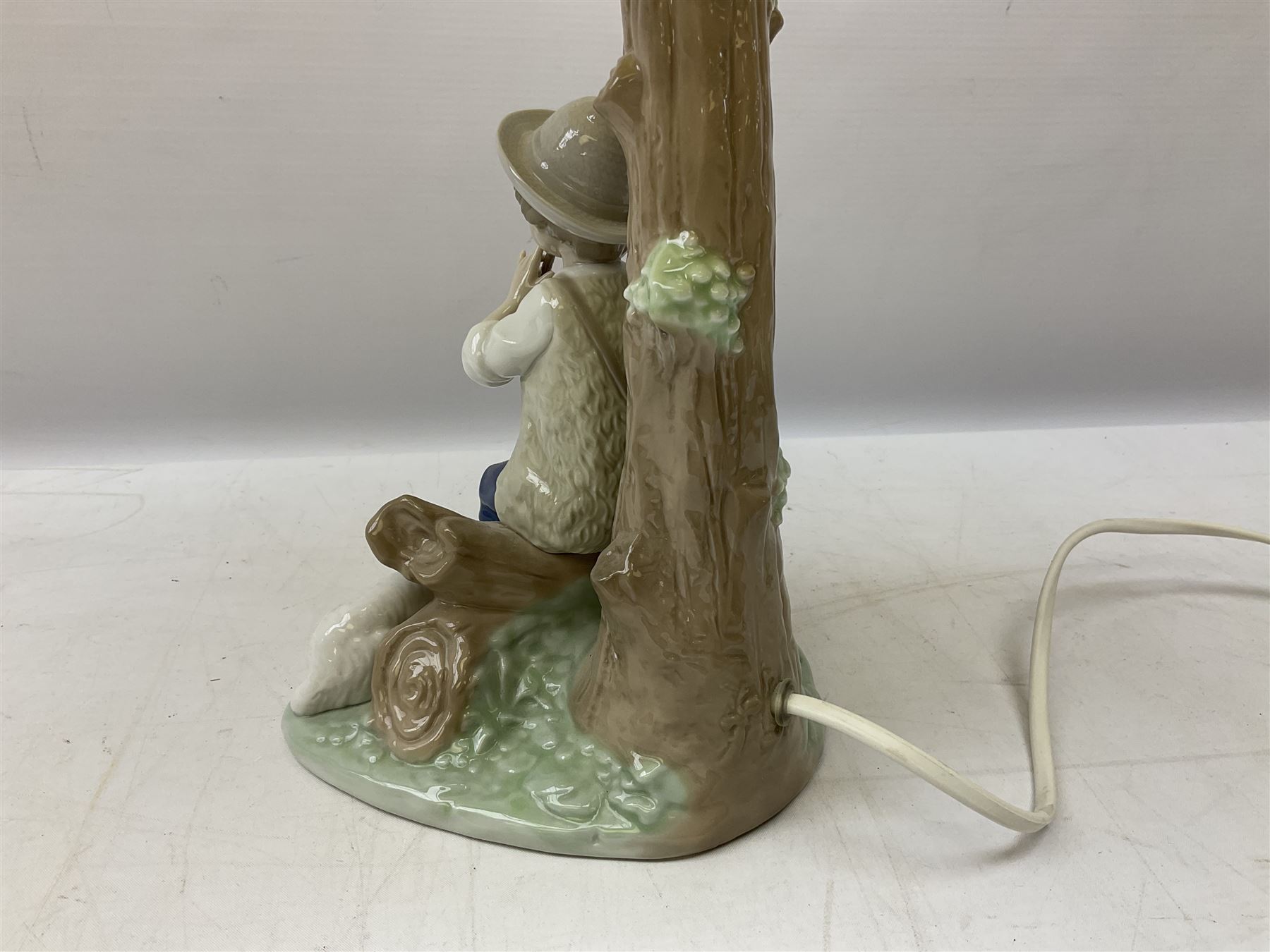 Nao table lamp modelled as a boy playing the flute upon logs with two recumbent sheep at his feet, the stem modelled as a tree trunk, with fabric shade, with printed mark beneath, H46cm