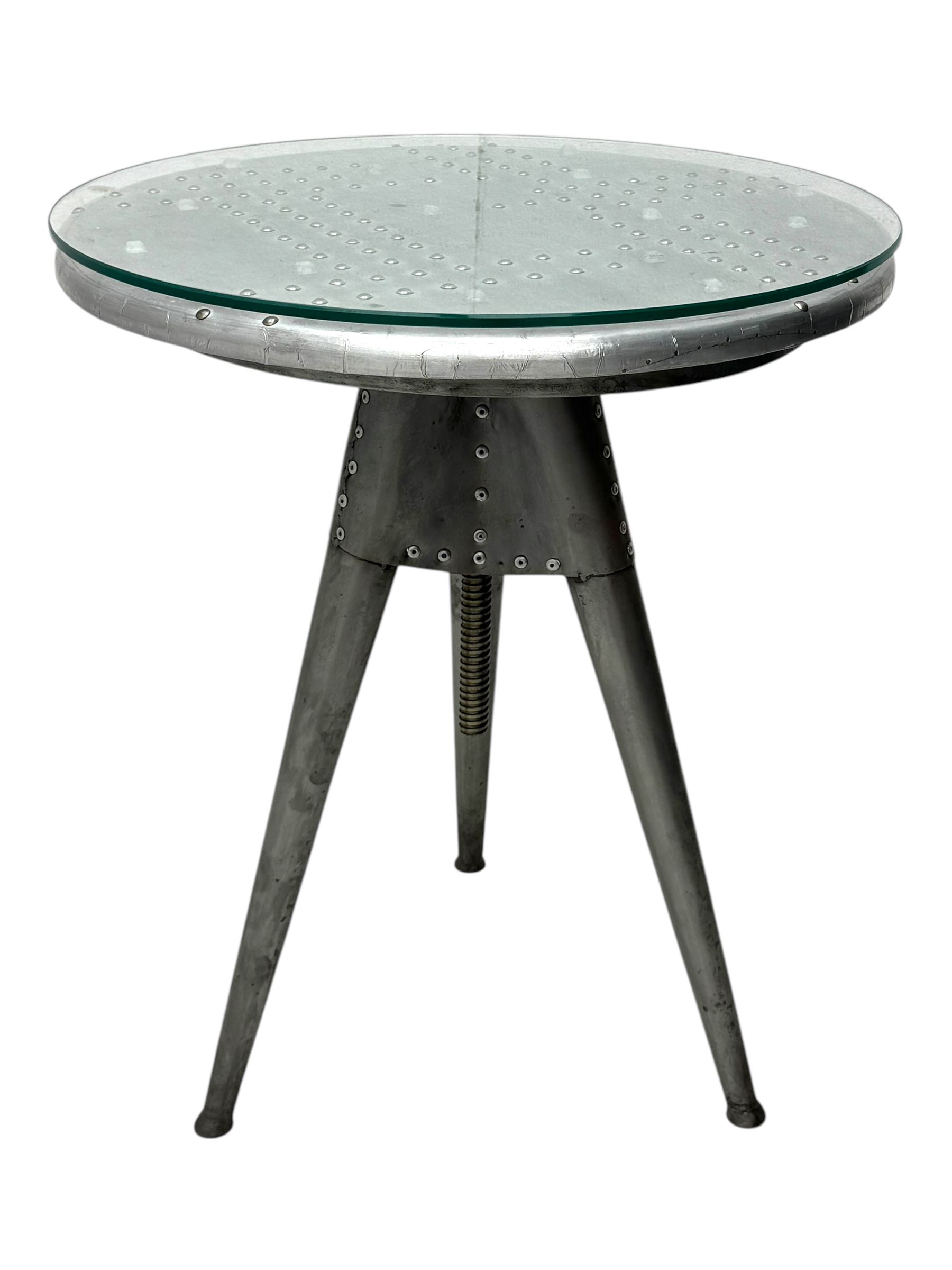Contemporary industrial design riveted metal table, by repute fashioned from salvaged aeroplane parts, with a circular glass top on a tripod base, accompanied by three adjustable stools upholstered in burgundy