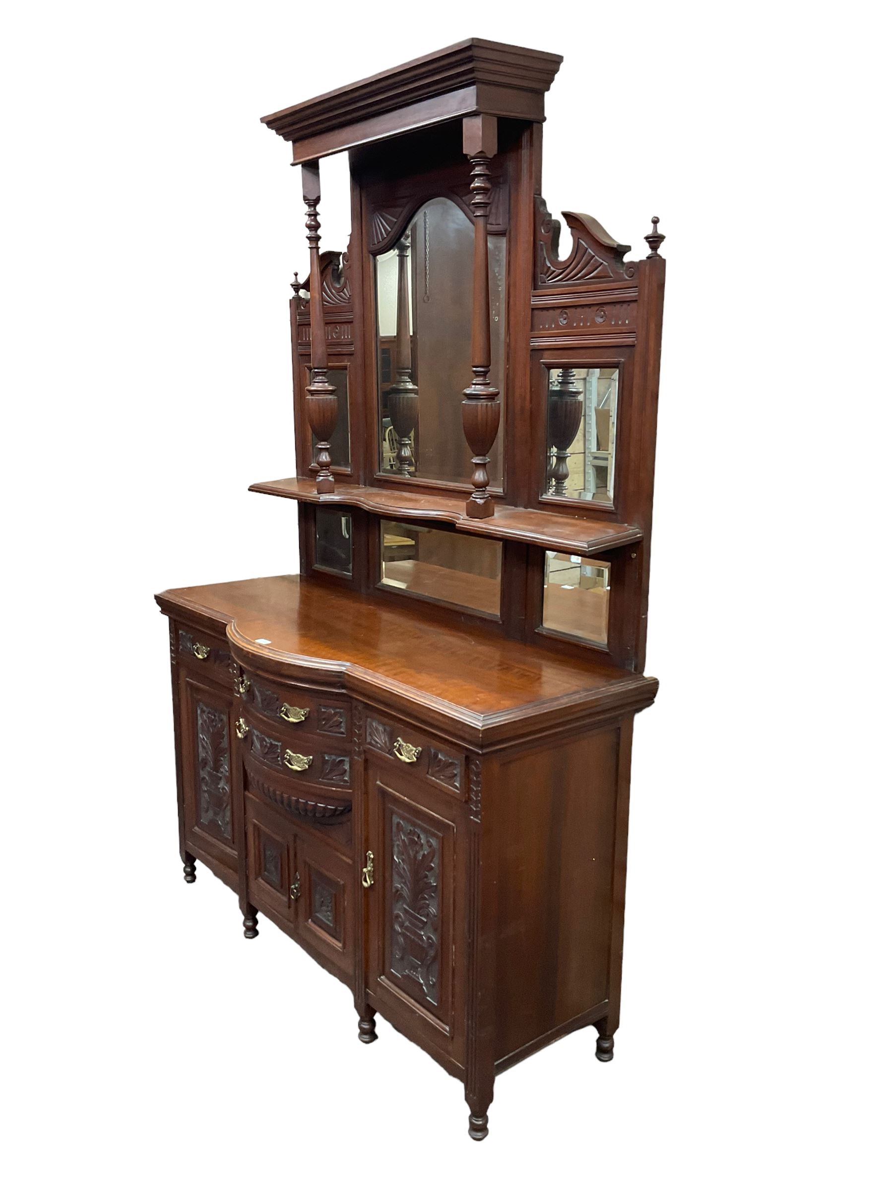 Edwardian walnut sideboard, projecting cornice supported by turned supports, the back fitted with three bevelled mirrors beneath fluted and flowerhead carved friezes, the lower section with four drawers and four panelled cupboard doors carved with foliate and urn designs, fluted uprights on turned feet
