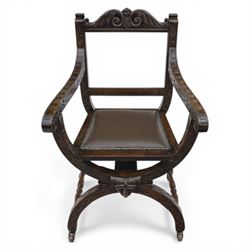 Late 20th century stained beech Savonarola chair, the carved crest rail and open arms above an upholstered leather seat, raised on an X framed base with carved foliate and mask decoration, joined by turned stretcher and ending in castors