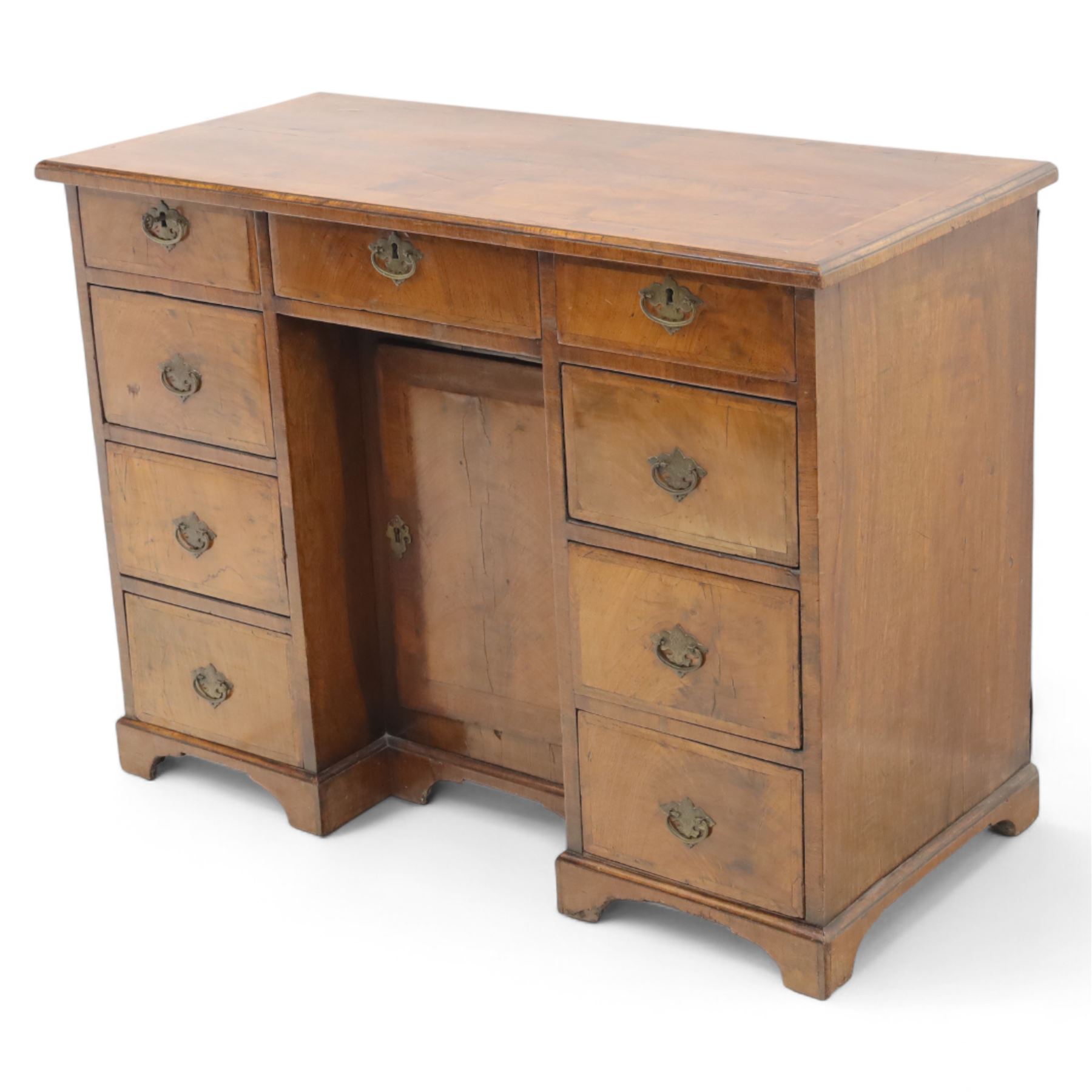 Early 20th-century Georgian-style walnut kneehole desk, the rectangular top with feather cross-banded border, above an arrangement of eight short drawers flanking a central cupboard door, fitted with brass backplate handles, raised on bracket feet