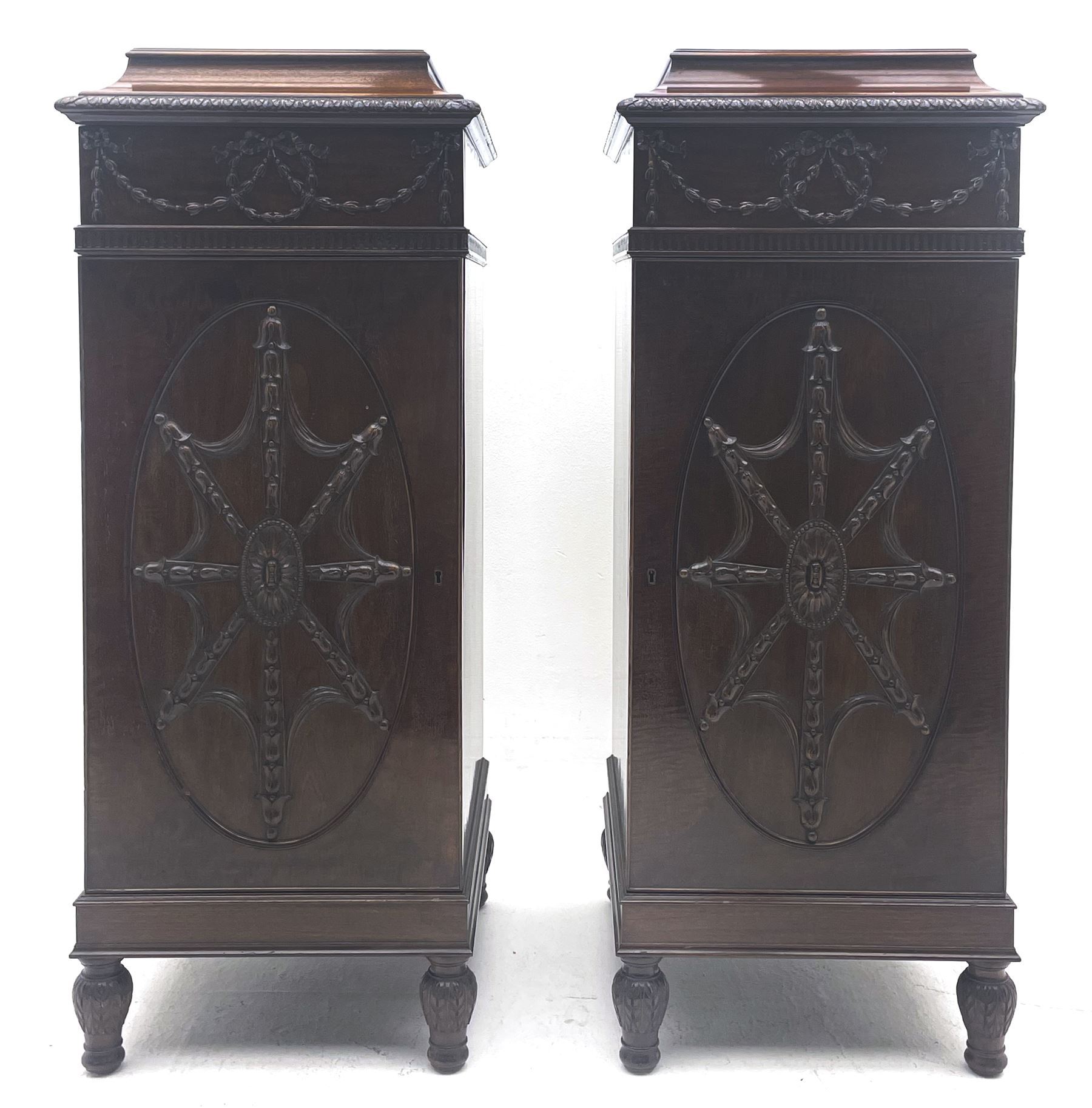 Pair early 20th century Hepplewhite style mahogany pedestal cabinets, cavetto moulded top with foliate carved edge over secret frieze drawer with applied trailing floral and ribbon decoration, each with button release underneath, oval panelled door with trailing harebell and central flower head motif decoration enclosing shelves, fitted cutlery drawer and lined cellarette drawer, on turned acanthus carved feet