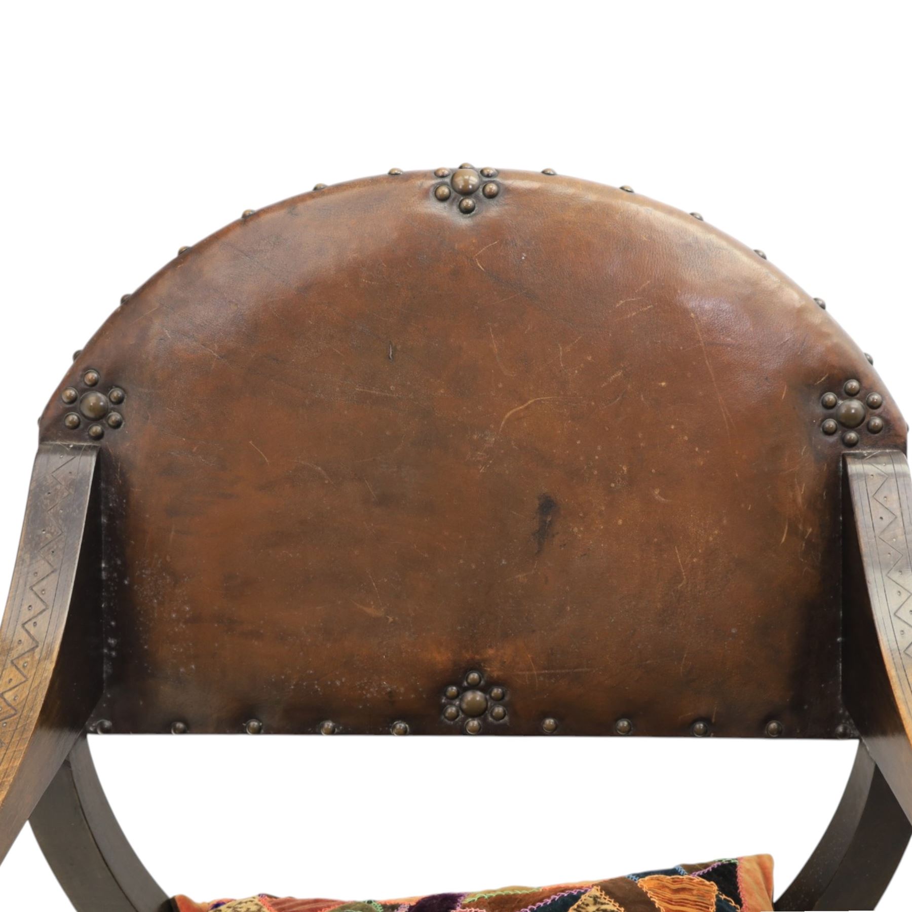 Early 20th century mahogany Savonarola chair, arched leather back with brass studwork, curved open arms with incised geometric decoration, loose patchwork velvet cushion seat, X-frame supports united by circular side rails, on scrolled feet