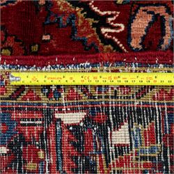 North West Persian Ahar hand-knotted carpet, madder red ground, with a large central shaped floral medallion with pendants, surrounded by angular scrolling foliate motifs incorporating rosettes, palmettes and stylised flowerheads, enclosed by ivory spandrels and a dark navy border with repeating geometric floral motifs and meandering vines, flanked by narrow patterned guard stripes, with cotton fringes