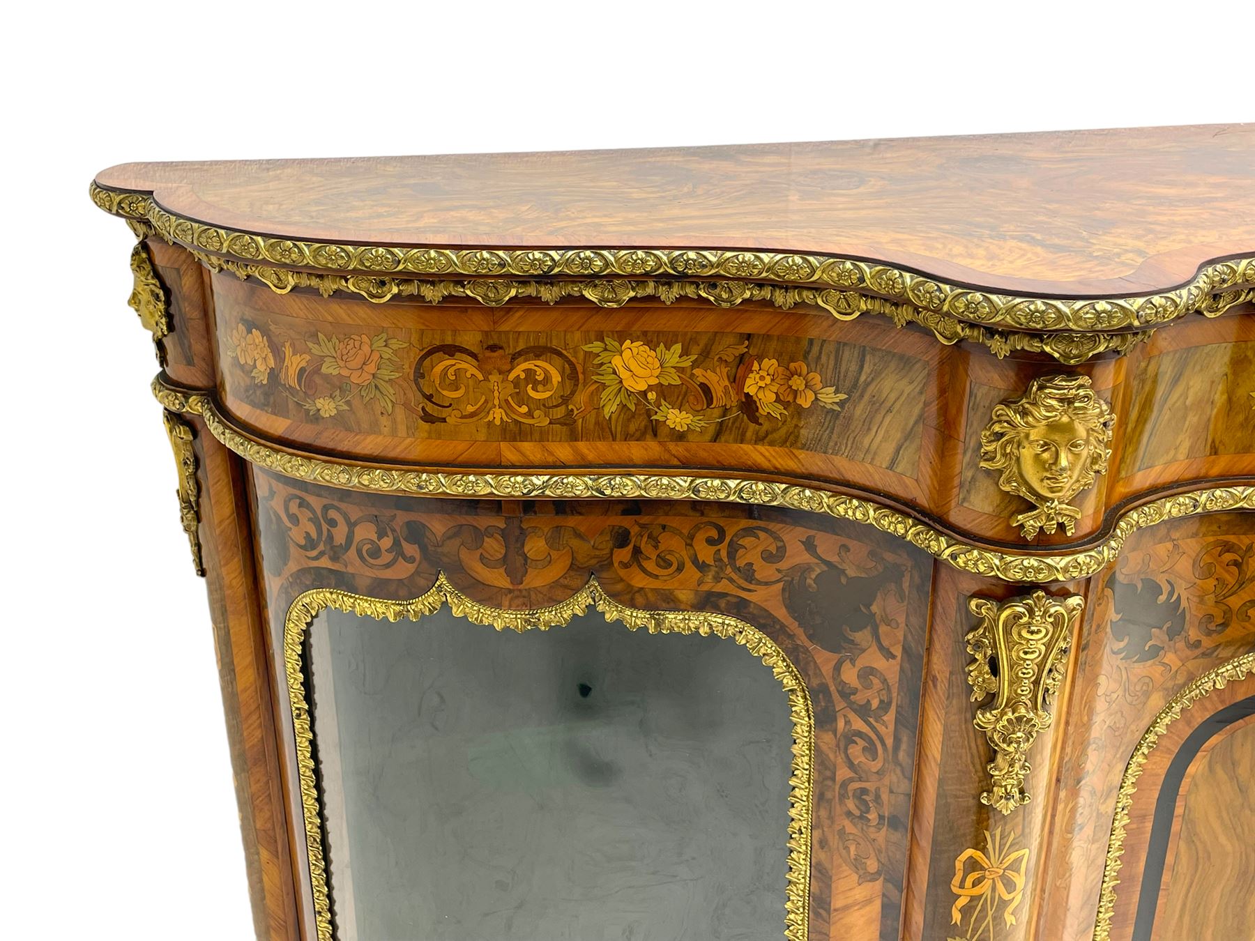 Victorian design inlaid figured walnut credenza sideboard, shaped form with crossbanded and book-matched top, the frieze rails inlaid with foliage scrolls and flower heads, central panelled door inlaid with floral urn flanked by two glazed doors, on shaped plinth base inlaid with foliate decoration, mounted by cast gilt metal masks and floral egg and dart beading 