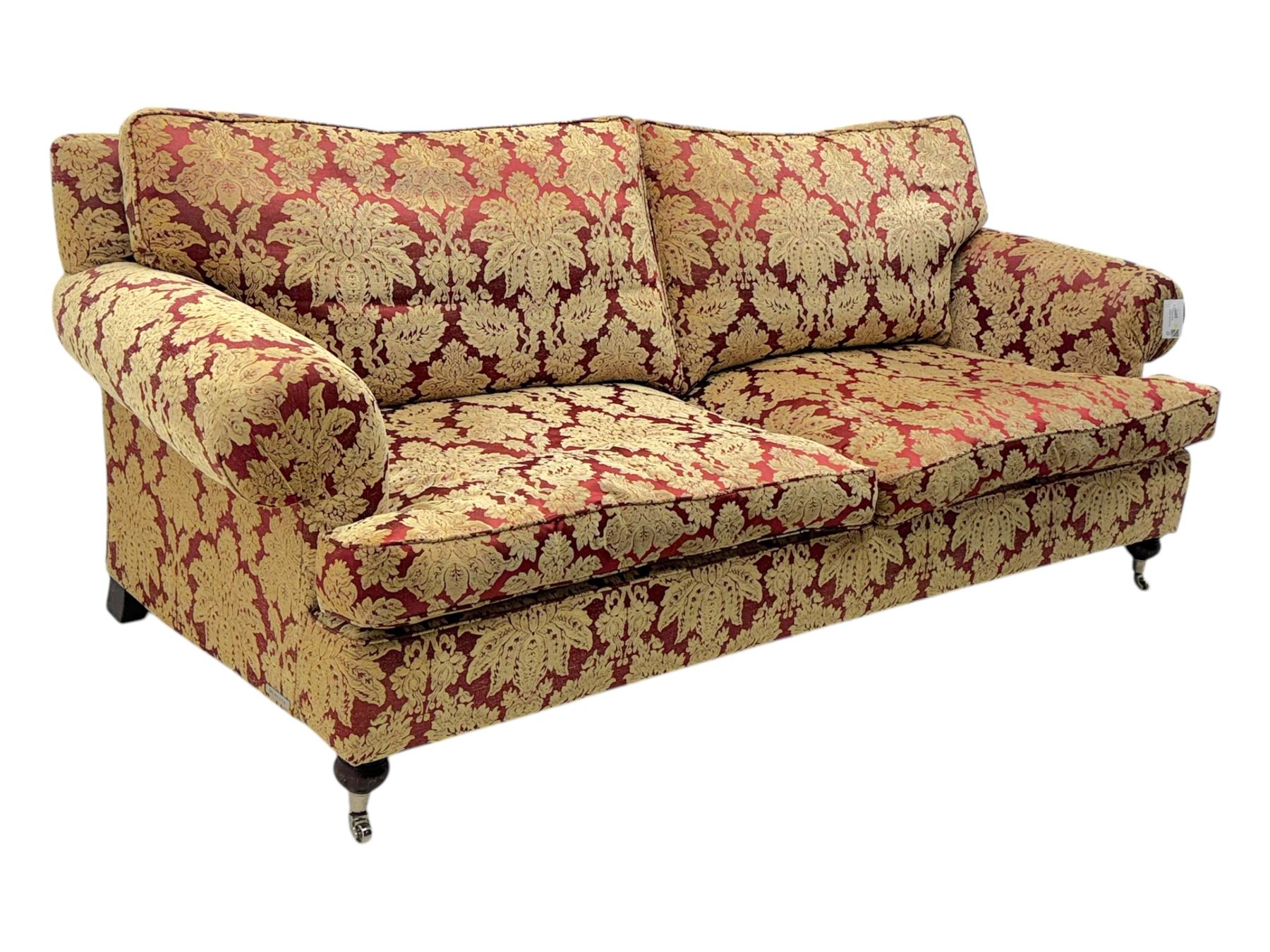 Duresta - pair of Grande three-seat sofas, traditional shaped with rolled arms and upholstered loose cushions, in red ground fabric with raised gold foliate pattern, on turned feet with brass castors 
