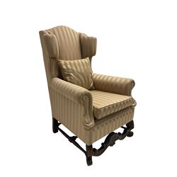 William and Mary style wingback armchair, upholstered in gold striped fabric, on walnut scroll carved supports joined by scroll and foliate carved middle rail, turned H-shaped stretchers, on castors