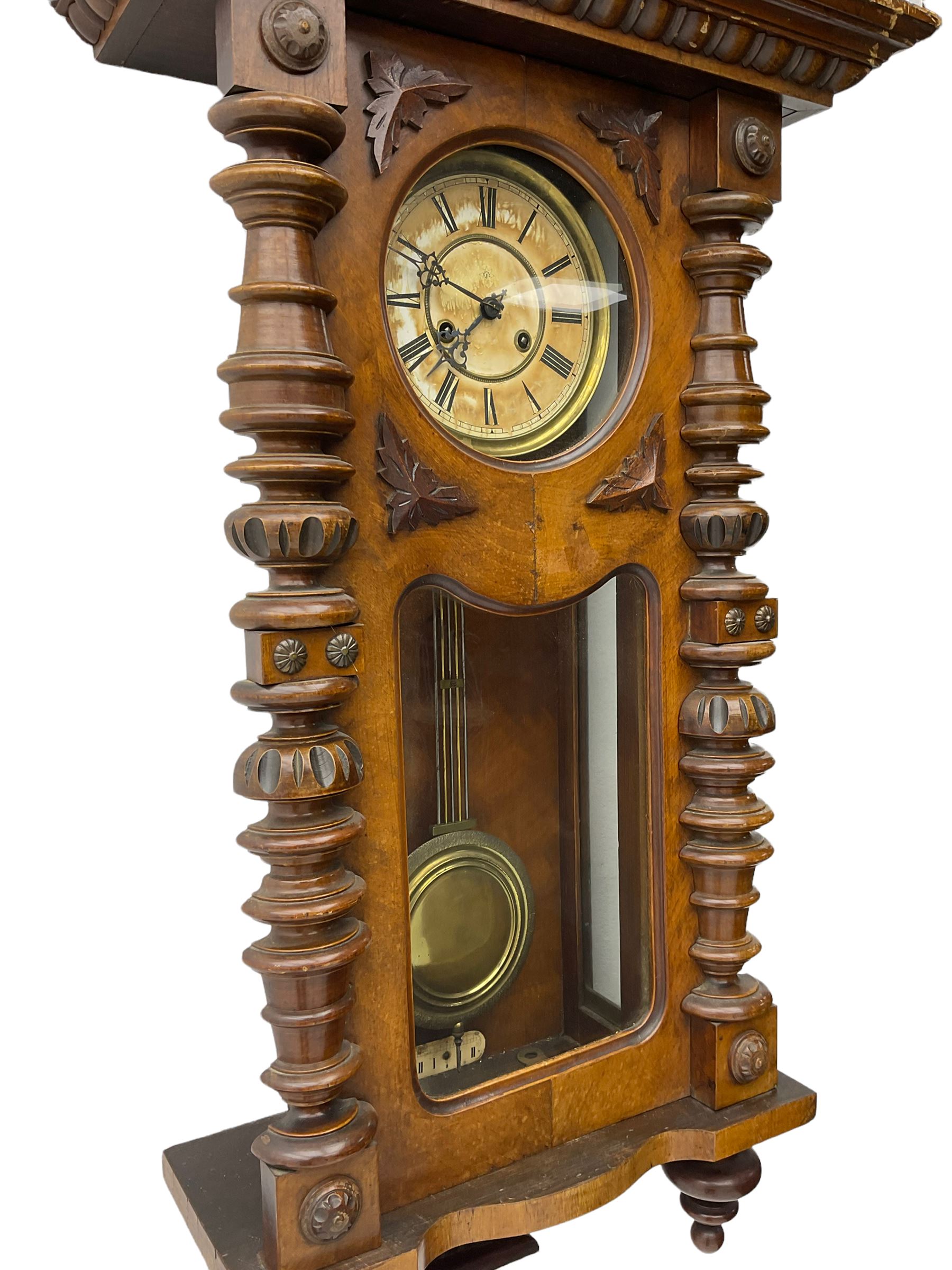 1890’s German striking wall clock in a walnut case, striking the hours and half hours on a coiled gong, eight-day Juhngans movement with a two-part dial, Roman numerals, gothic steel hands and visible gridiron pendulum, case with turned pilasters, applied carving and glazed full length door.

