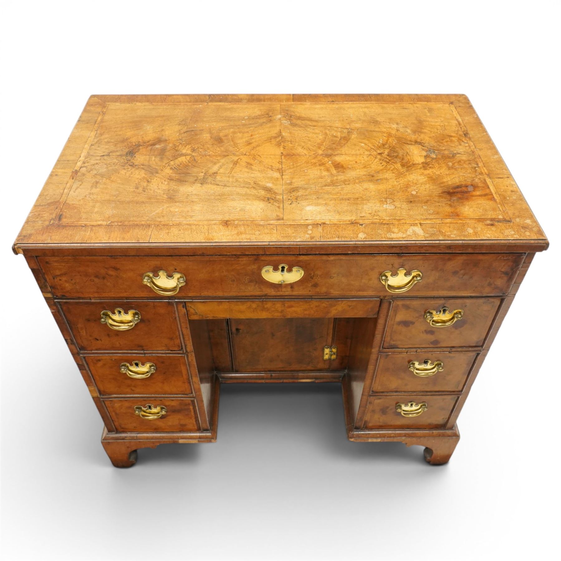 George II figured walnut kneehole desk, the rectangular book-matched veneer top with feather and cross-banding, fitted with single frieze drawer over a short drawer and central recessed cupboard, flanked by three drawers to each side, lower moulded edge on bracket feet
