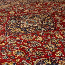 Central Persian Kashan hand-knotted carpet, crimson red central field with central floral medallion with floral vine-work and palmettes, within dense floral spandrels and a wide navy blue border decorated with continuous foliate motifs and multiple guard borders, with cotton fringe ends  