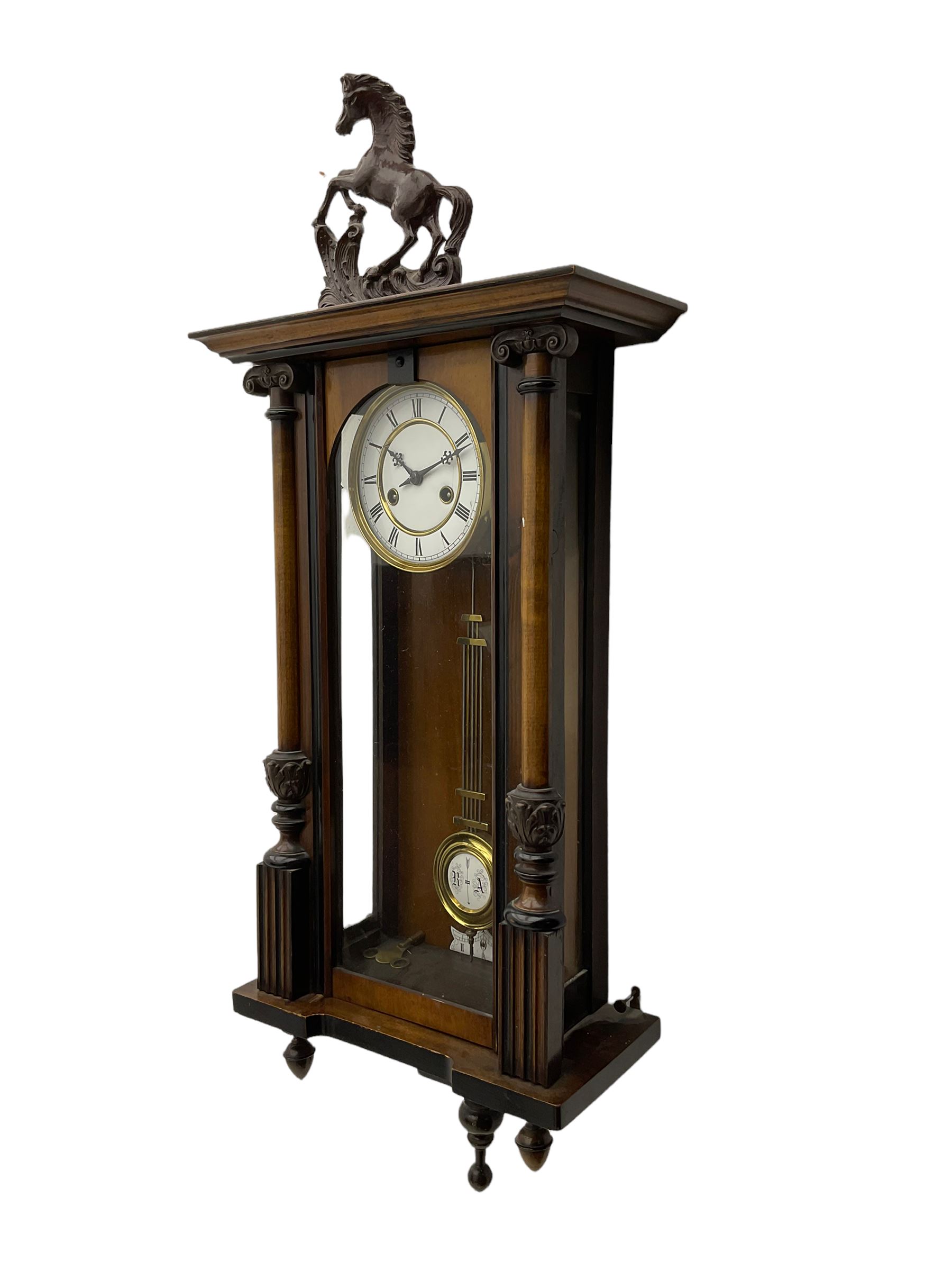 A late 19th century German spring driven 'Vienna' wall clock with a fully glazed door flanked by turned columns with ionic capitals and carving, replacement pediment depicting a prancing horse, with a two-piece enamel dial, Roman numerals, minute track and gothic steel hands, 8-day movement striking the hours and half hours on a gong,  with a gridiron pendulum and beat plate. With key.


