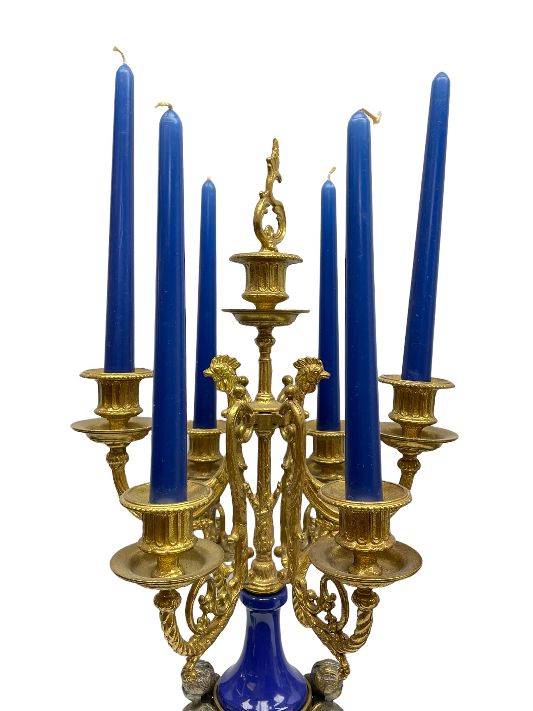 A 20th century continental gilt metal Lyre mantle clock with a pair of matching six light candelabra, spring driven movement housed in a gilt drum case on a brass and cobalt blue base with paw feet, two bronzed metal figures of mythological cherubs with animal legs and cloven hooves supporting a festooned garland swag, eight-day twin barrel striking movement with a floating lever balance escapement, striking the hours and half-hours on two bells, white enamel dial with roman numerals, minute track and pierced steel hands, dial inscribed “Imperial”. 