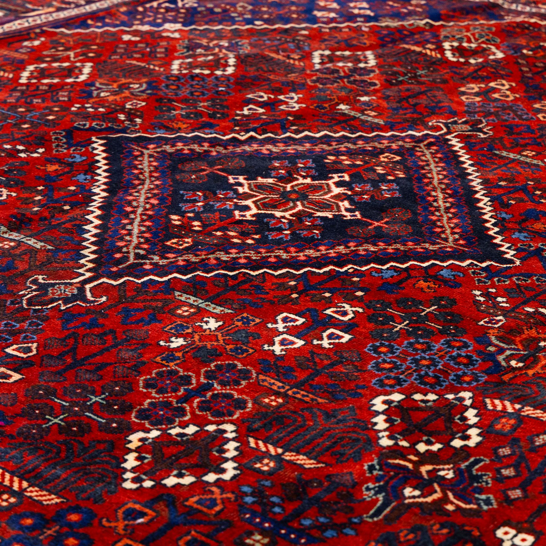 Central Persian Joshagen hand-knotted carpet, madder-red field with a central stepped lozenge medallion and densely filled with stylized geometric floral and tree motifs arranged in a repeating lattice enclosed by indigo spandrels and a broad navy border of angular palmette and vine ornament, flanked by multiple guard stripes, cotton fringe ends 