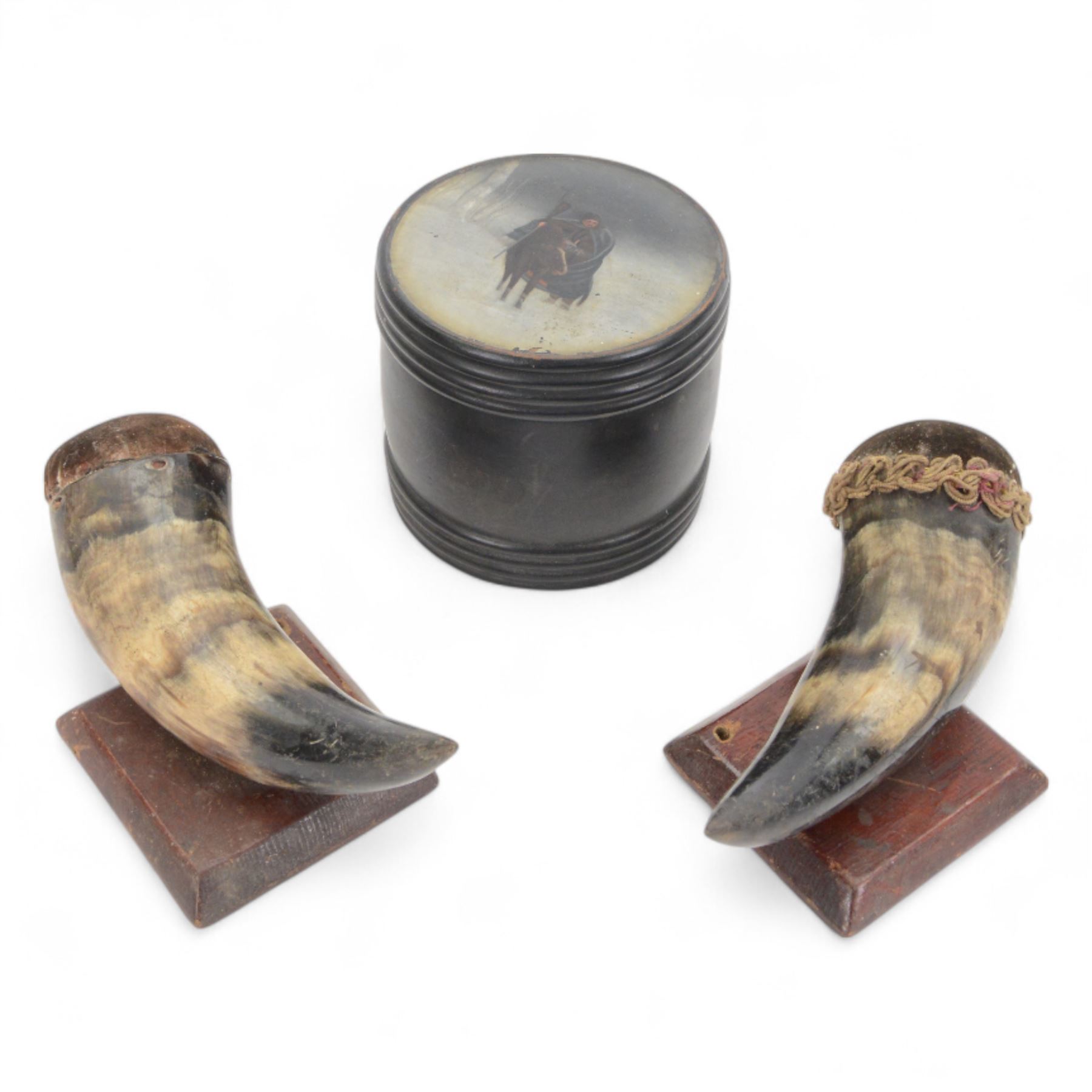 19th Century Russian lacquered tobacco box the painted panel top depicting a man on horseback in the snow, together with two horn pin cushions upon wooden plinths 