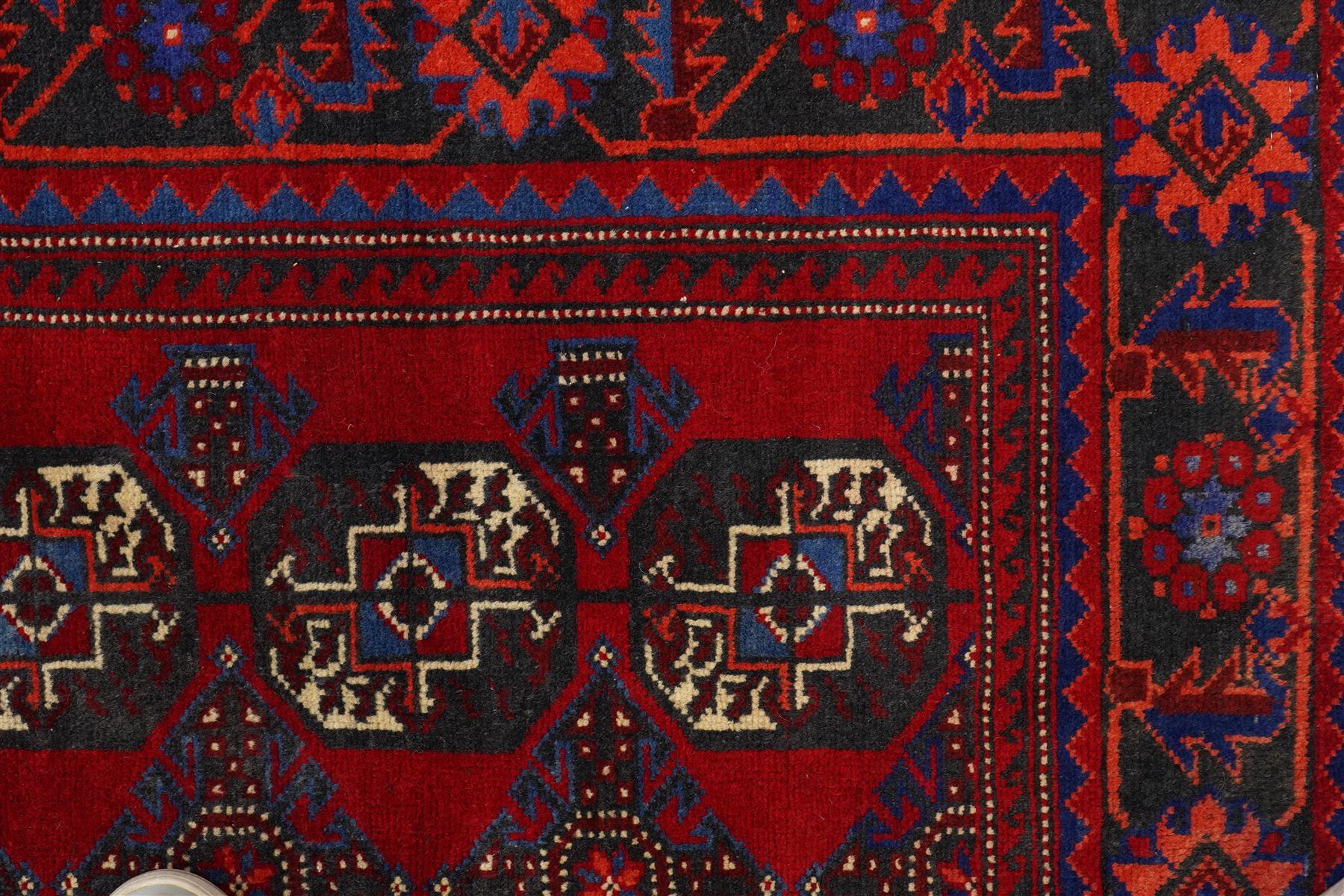 North East Persian Balochi hand-knotted runner, crimson field decorated with three vertical columns of repeating octagonal guls, each enclosing angular geometric motifs in deep indigo, ivory, and soft brown tones, guls linked by stepped diamond and cruciform devices, main border of stylized geometric floral motifs and angular vines, flanked by multiple guard borders, with cotton fringe ends 