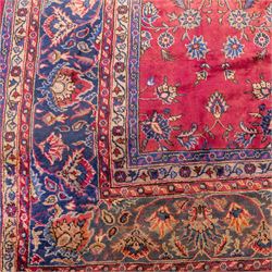 Large Persian-style mahal-type hand-knotted carpet, of rectangular form with a deep crimson field decorated with an all-over arrangement of stylized floral sprays and palmettes, with a central subtle medallion composition enclosed by a dark navy main border of scrolling floral motifs between narrow guard stripes with fringe ends 