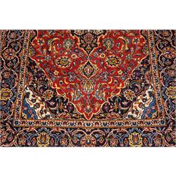 Central Persian Kashan hand-knotted rug, madder red field, with central lobed medallion with pendants, issuing an all-over design of scrolling vine, palmettes, and floral sprays, with conforming spandrels, enclosed by a broad indigo palmette and vine border framed by guard stripes in a palette of navy, blue and ivory tones, with cotton fringe ends 