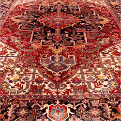 North West Persian Heriz hand-knotted carpet, madder red field with large central stepped medallion with angular floral and geometric designs, surrounded by stylized palmettes, leaves and tribal motifs with indigo corner spandrels, enclosed by a broad navy border of geometric floral devices between multiple guard stripes, with cotton fringe ends 