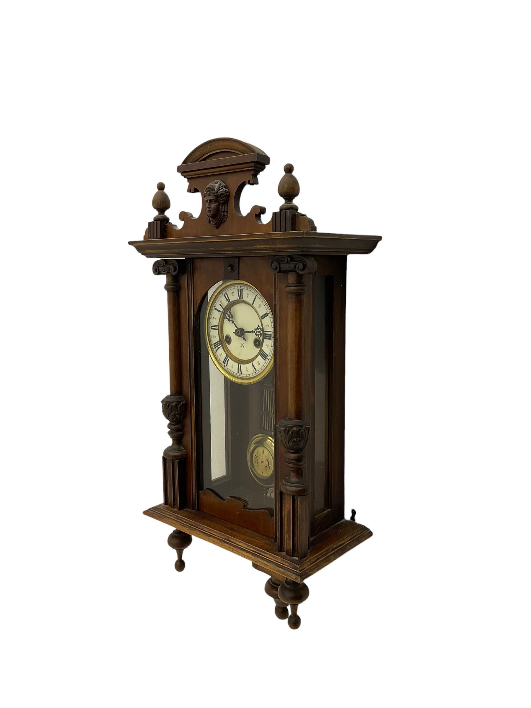 An early 20th century German wall clock with an 8-day HAC spring driven movement striking the hours on a coiled gong, in a mahogany case with a shaped pediment and turned columns flanking a full-length glazed door, with a two-part dial enamel with Roman numerals, minute track and pierced gothic designed hands, gridiron pendulum and beat plate. With Key.





