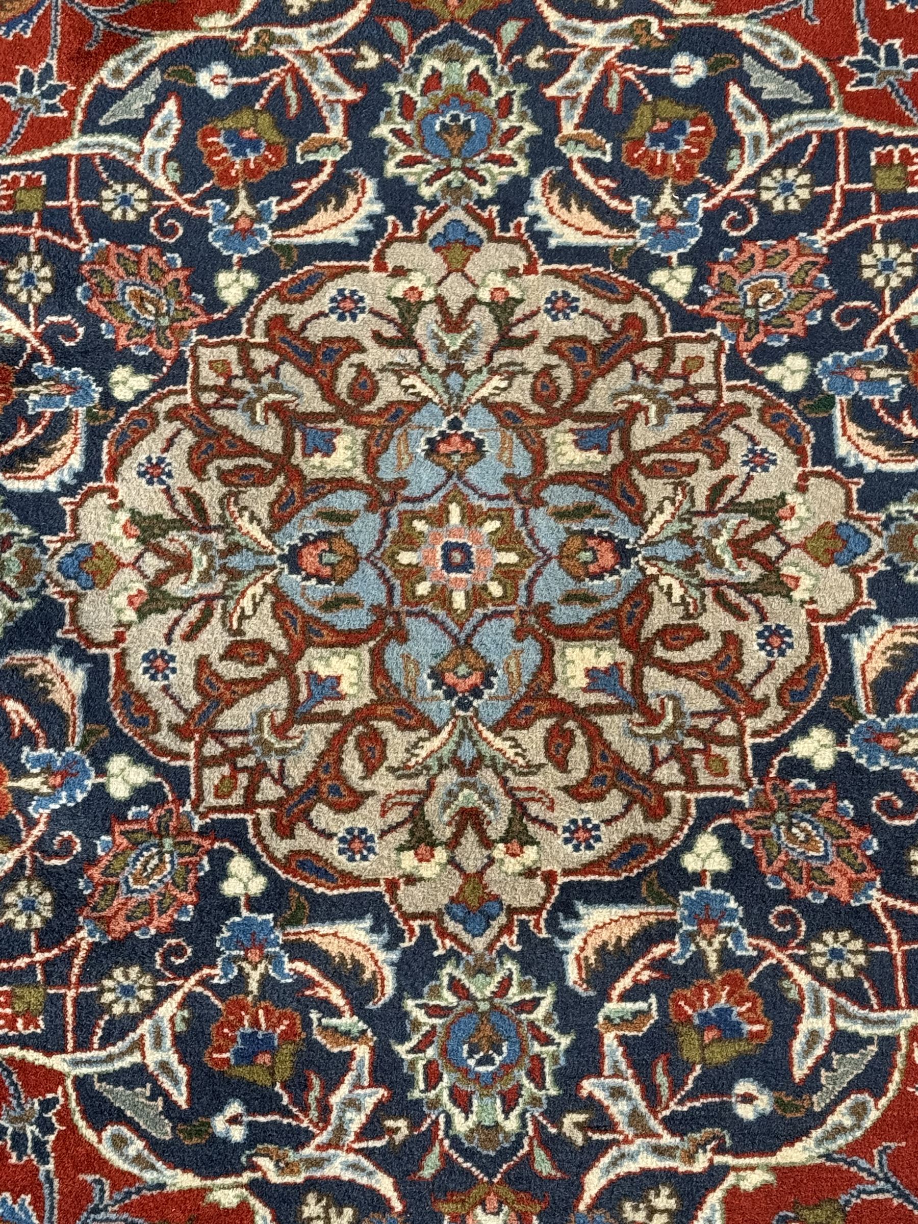 Central Persian Kashan carpet, the red field centred by a large multi-lobed floral medallion, flanked by corner spandrels in navy, enclosed by multiple scrolling floral borders, fringed to two ends