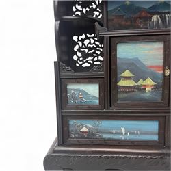 Early 20th century Japanese Meiji style carved hardwood cabinet, of symmetrical tiered form with pierced foliate fretwork panels and phoenix crest, fitted with sliding and hinged doors inset with hand-painted landscape panels depicting Mount Fuji, waterfalls, and lakeside dwellings, raised on shaped bracket supports