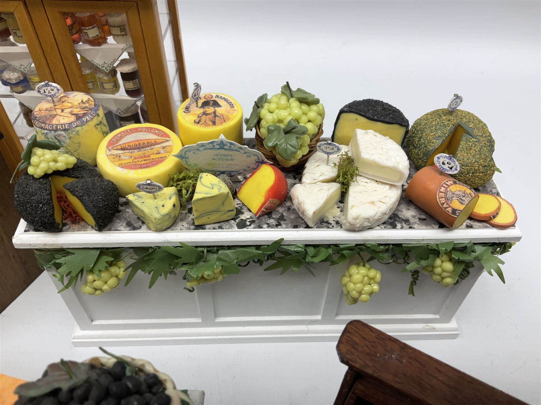 Collection of miniature dolls house grocery shop furniture and produce, comprising glazed corner counter with brass style cash register, shelved preserves unit, pies, cheese board and trays of meats, pair of white cheese counters, biscuit display with six tins, glazed preserves cabinet, pair of shop chairs and four further cheese boards complete with grapes and label flags