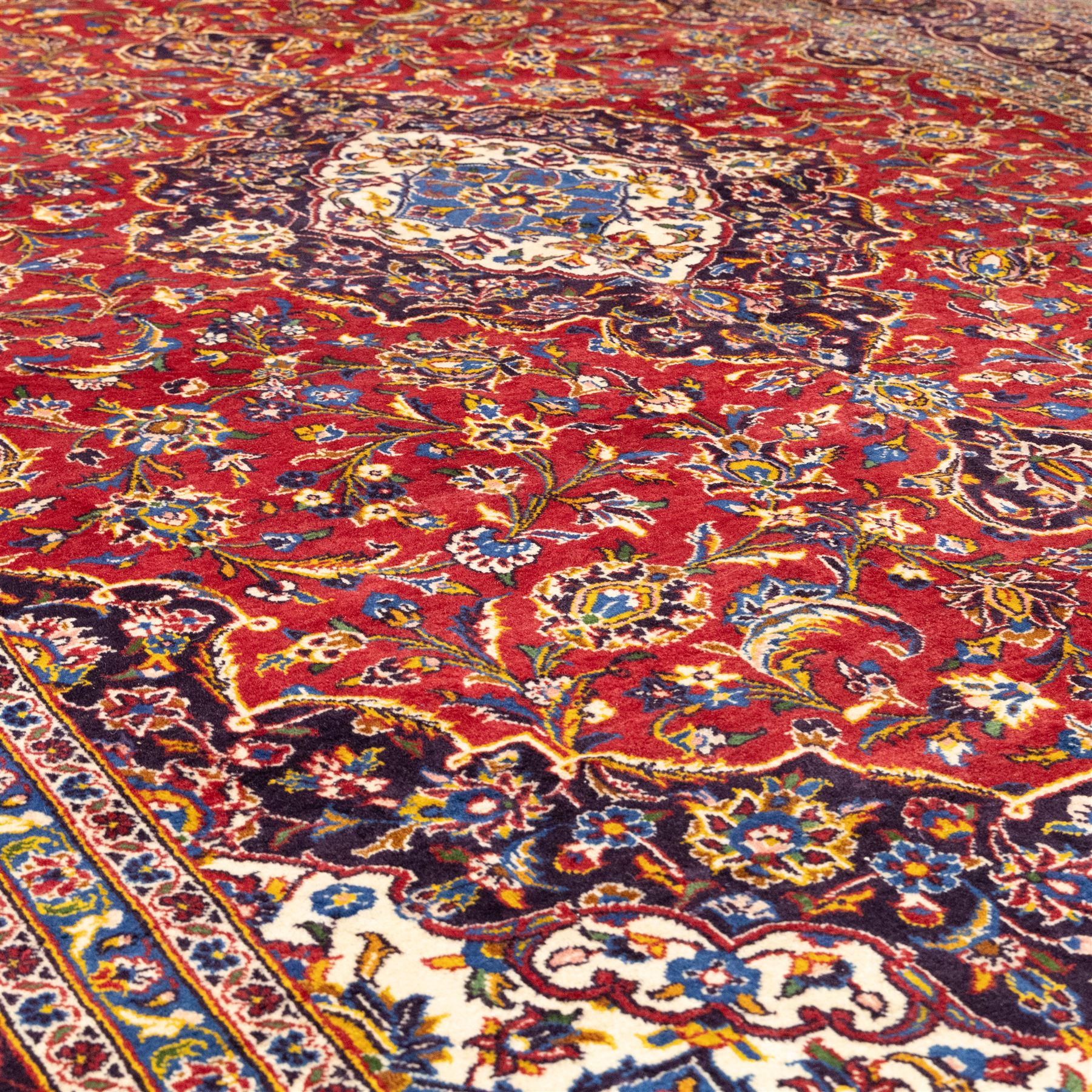 Central Persian Kashan hand-knotted carpet, crimson central field with lobbed medallion in navy blue and ivory ground, surrounded by a dense all-over floral design of scrolling vines, palmettes, and stylized blossoms, enclosed within a deep navy main border, flanked by multiple guard stripes with fringe ends 


